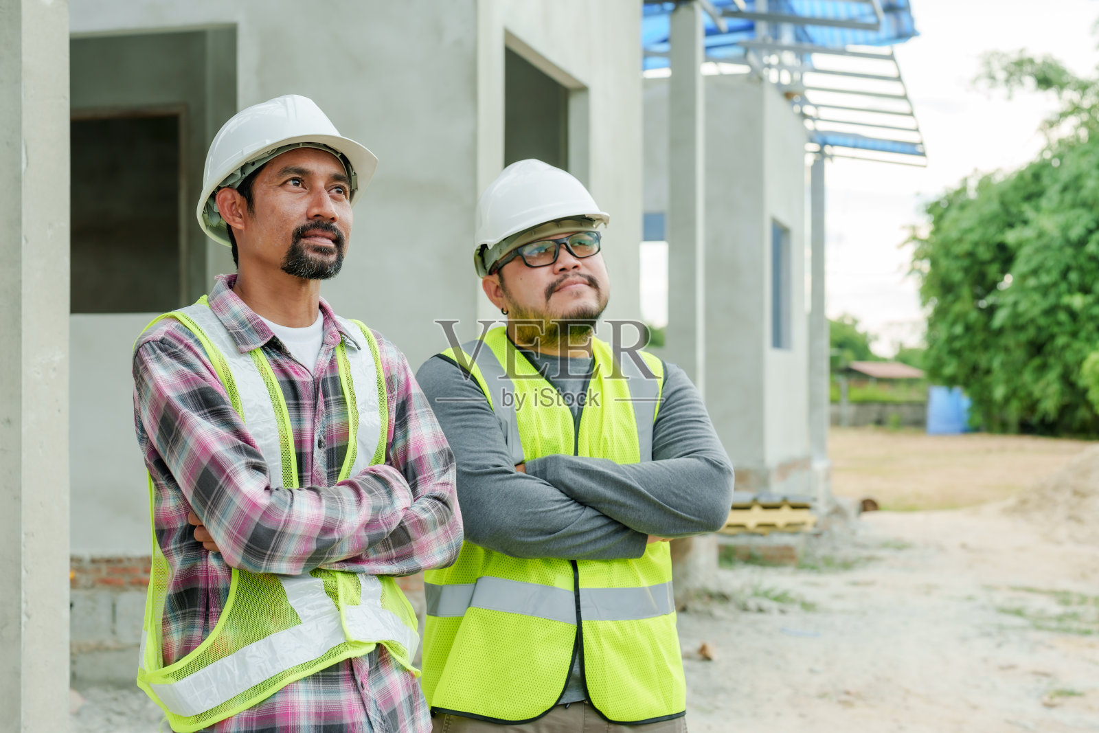 两名亚洲男性工程师，在施工区域戴着头盔保护头部，为了安全穿着全套服装，双臂交叉站立，抬头看着正在建造的房屋，站在建筑工人旁边。照片摄影图片
