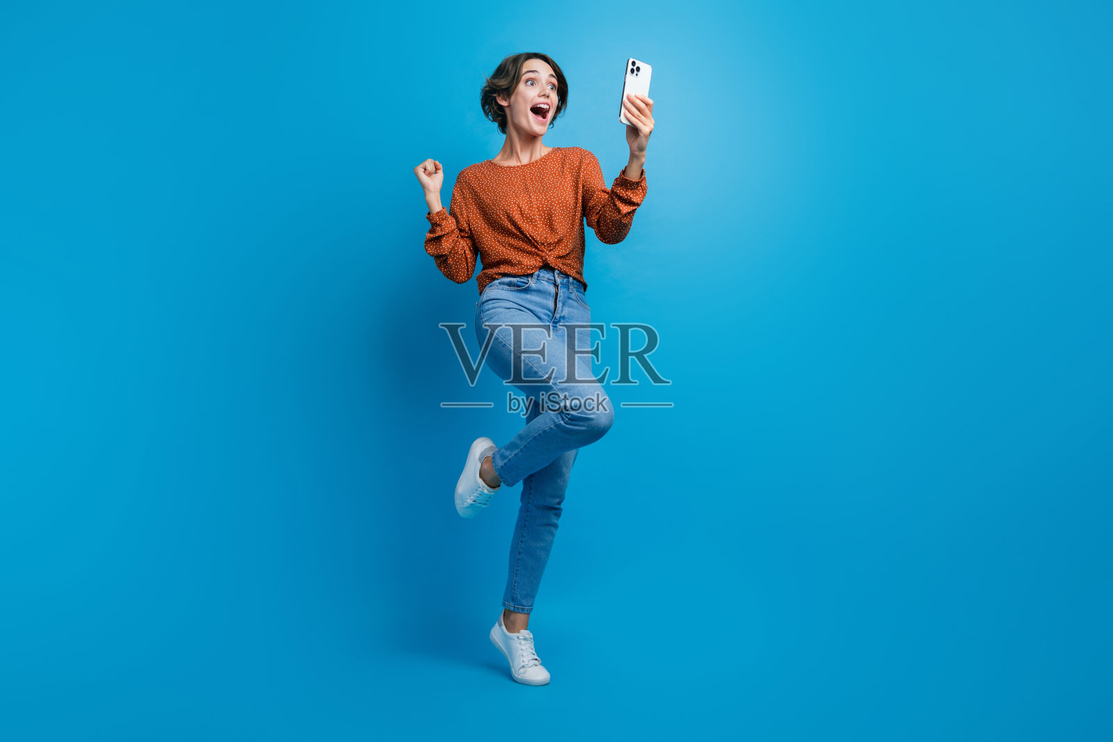 可爱的年轻女士庆祝大折扣店手持设备穿着时髦的棕色服装孤立在蓝色背景的全身照片照片摄影图片