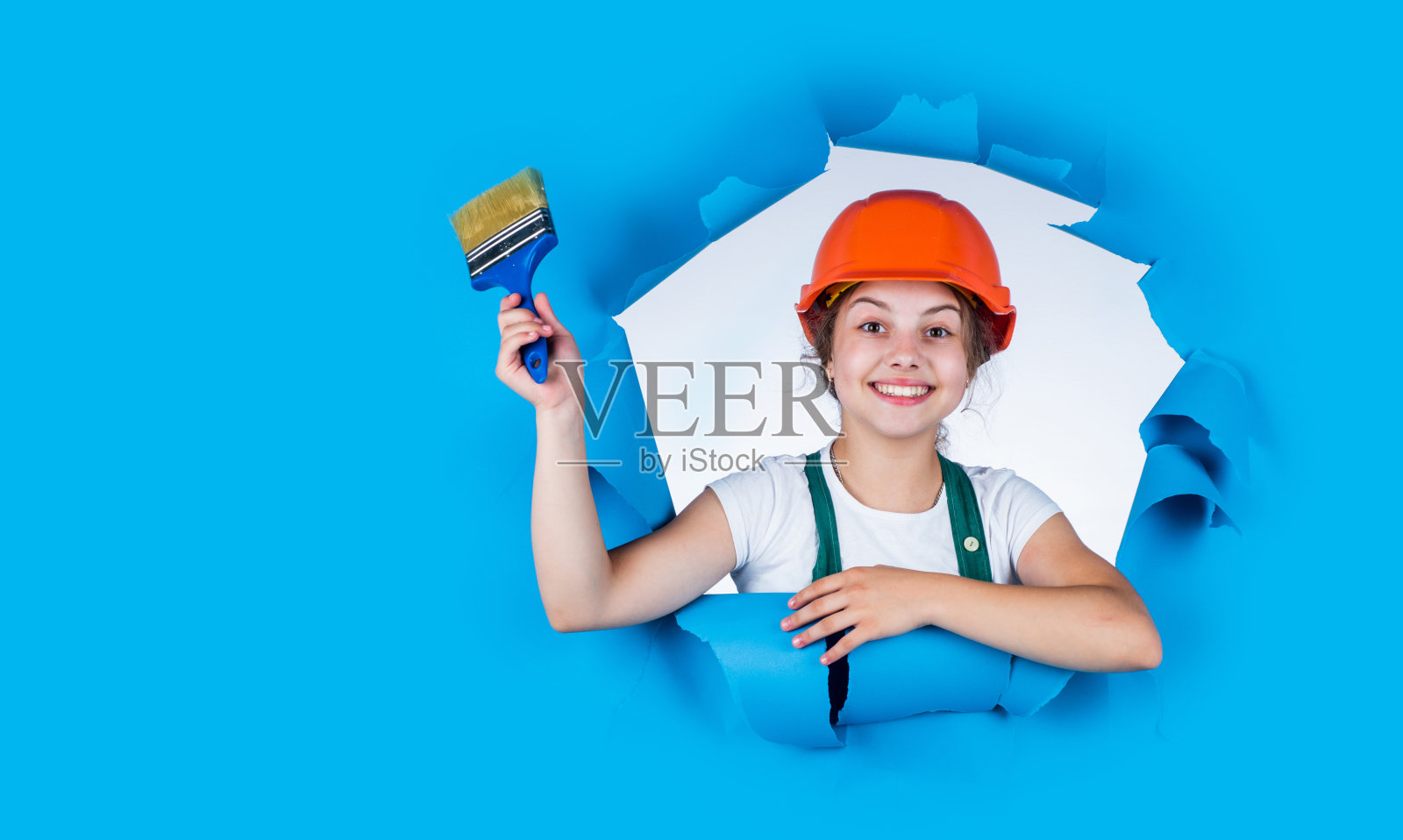 戴着防护头盔的女孩在粉刷墙壁。儿童的发展。建筑工女孩。快乐的童年。快乐的女孩戴着安全帽，手里拿着画笔。准备上色照片摄影图片