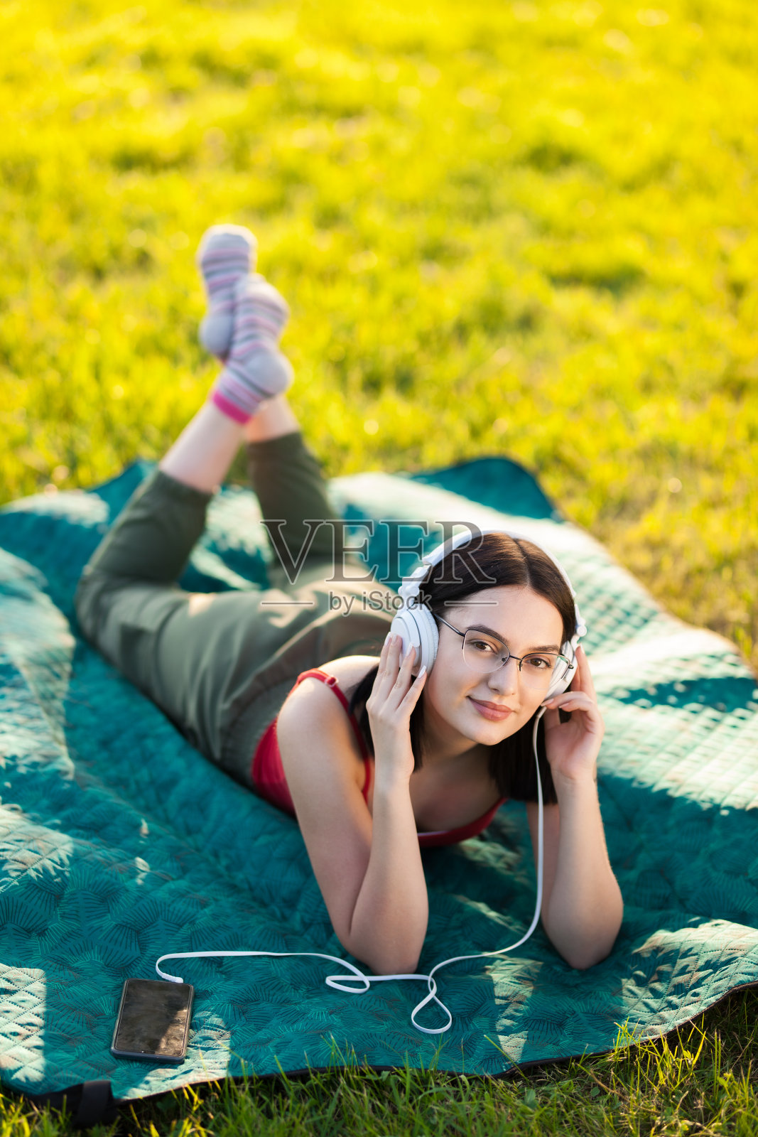 黑发女孩用智能手机戴着耳机听音乐。躺在绿色的草地上。照片摄影图片