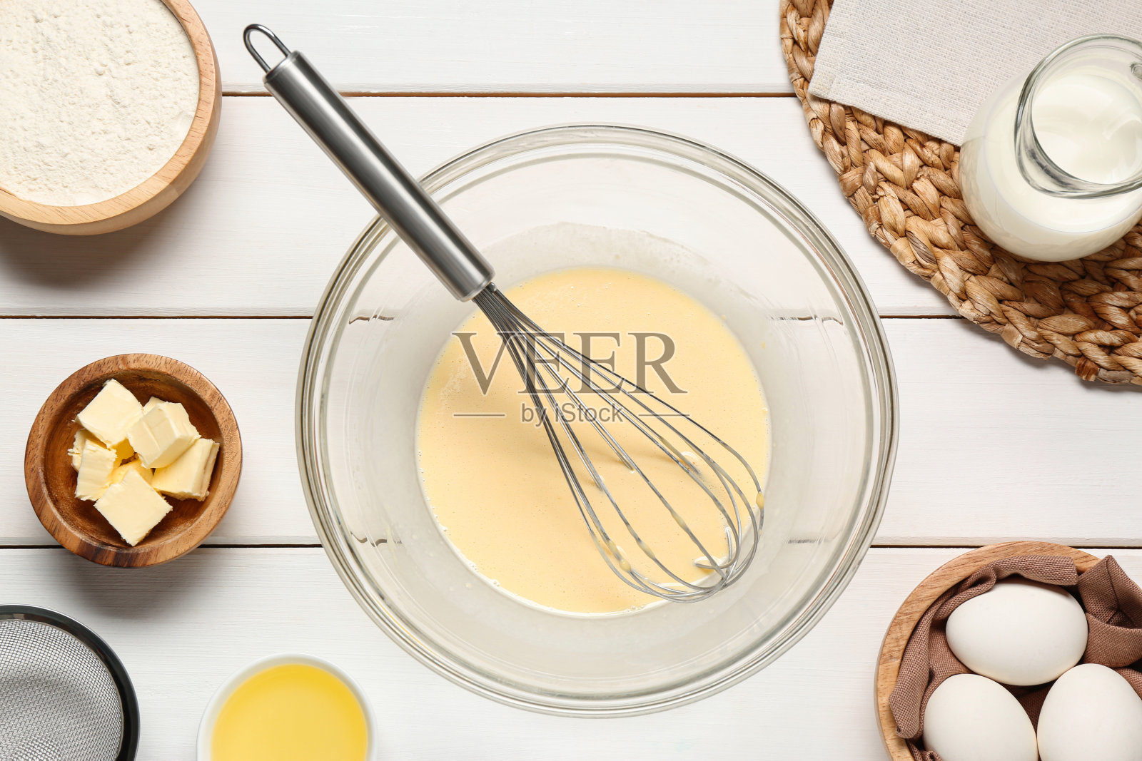 玻璃碗的可丽饼面糊与搅拌器和配料在白色木桌上，平放照片摄影图片