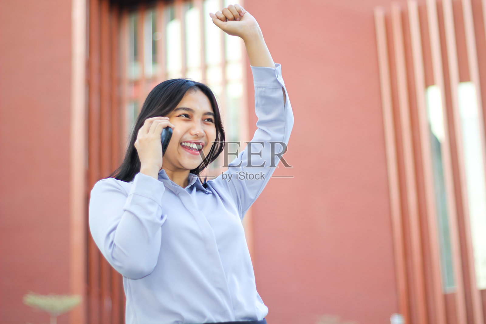 欢快的亚洲女商人打电话庆祝胜利，举起拳头说“是”，穿着正装，女会计站在红色的建筑物上用智能手机讲话照片摄影图片
