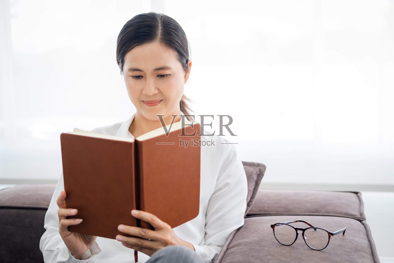 灰色舒适的沙发沙发和一个美丽的亚洲女人，看书，家的概念和舒适，文字复制空间的地方。照片摄影图片