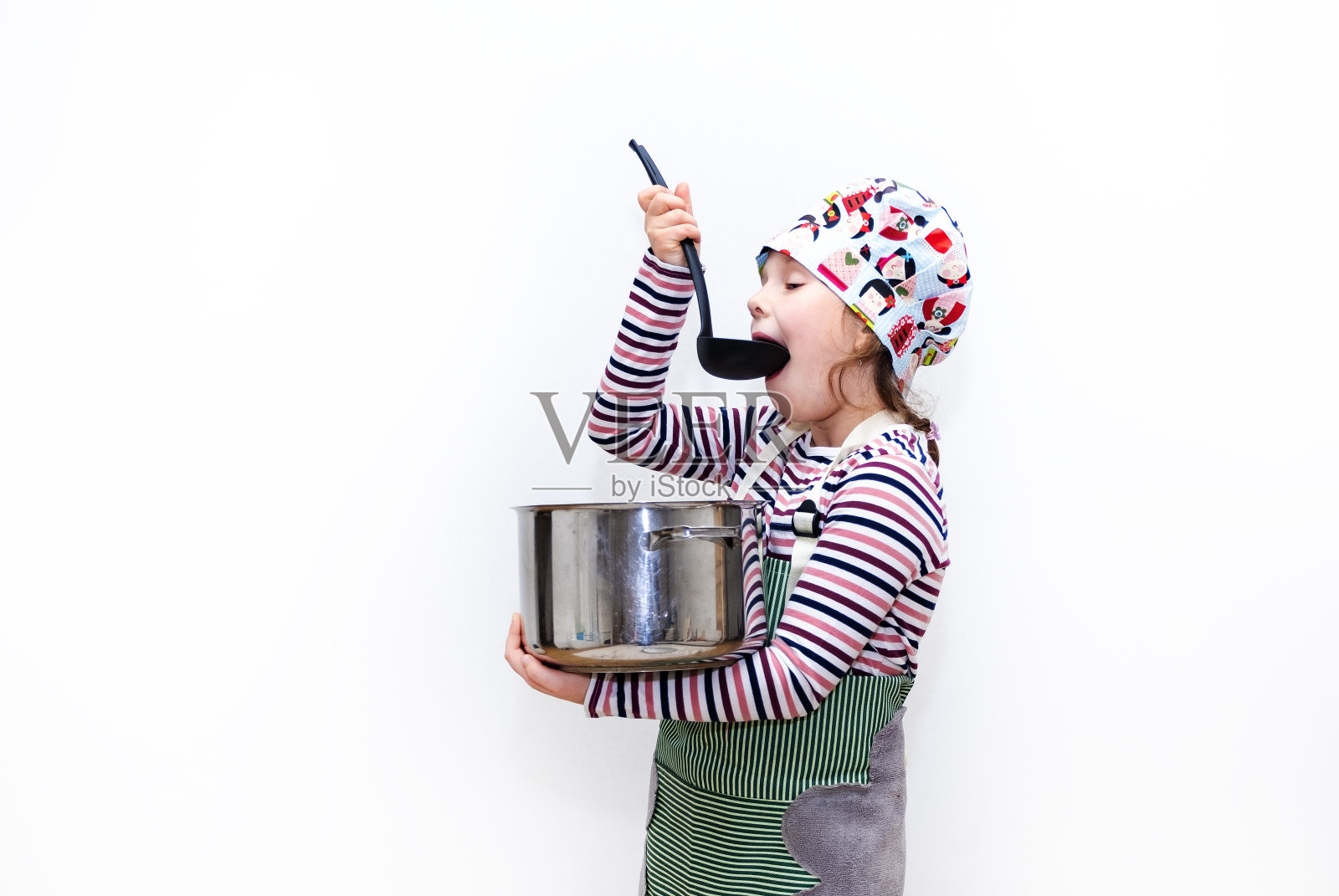 一个8岁的白人女孩，打扮成厨师，手里拿着一个勺子和一个大炖锅，品尝着食物。情感的脸。照片摄影图片