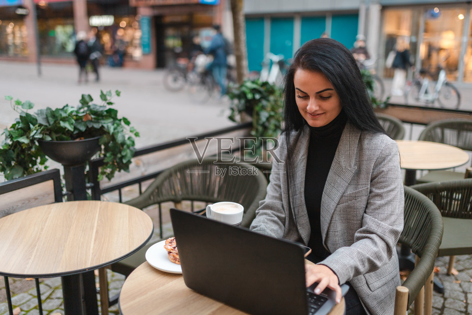 漂亮的年轻女商人坐在咖啡馆里一边工作一边端着一杯咖啡和一个肉桂圆面包照片摄影图片