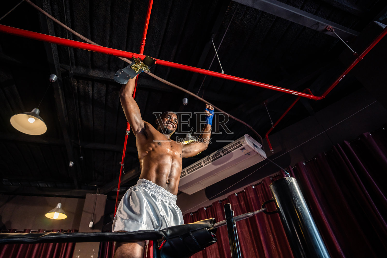 非洲肌肉拳击手在赢得比赛后在舞台上举起双臂。俊俏的男运动员拳手赤膊上阵，心情愉悦，兴奋不已，在赛场上的拳台上享受拳击运动。照片摄影图片