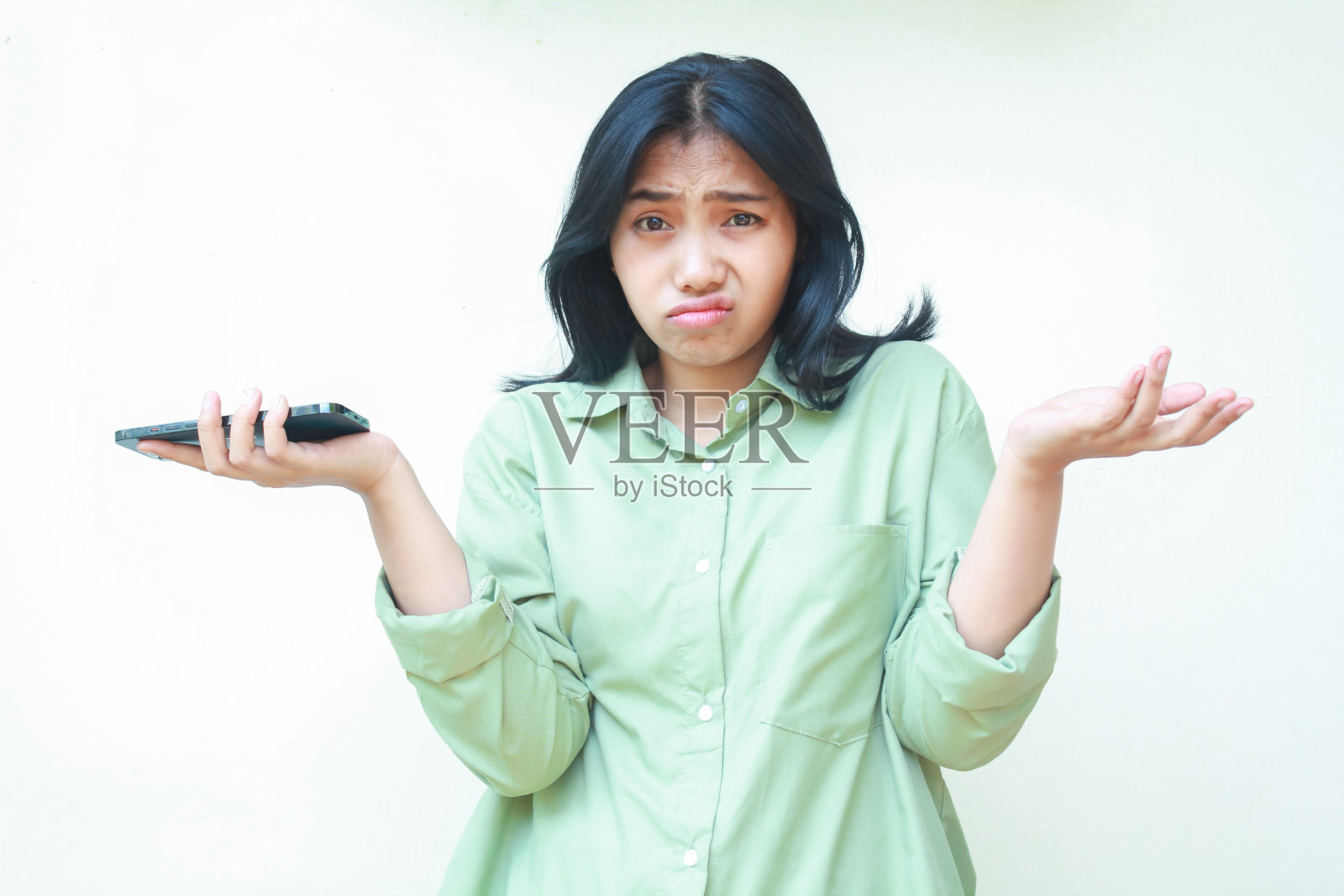 一名穿着绿色衬衫的困惑亚洲女子的肖像耸肩，手掌张开，拿着智能手机，看着相机，担心地站在孤立的背景上，考虑着手势照片摄影图片