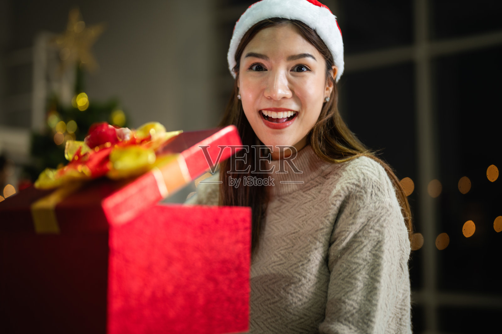 年轻的亚洲微笑的漂亮女人拿着红色的礼盒庆祝新年。圣诞礼物。女性穿着白色针织毛衣，头戴帽子，与圣诞树合影。冬季流行趋势。照片摄影图片