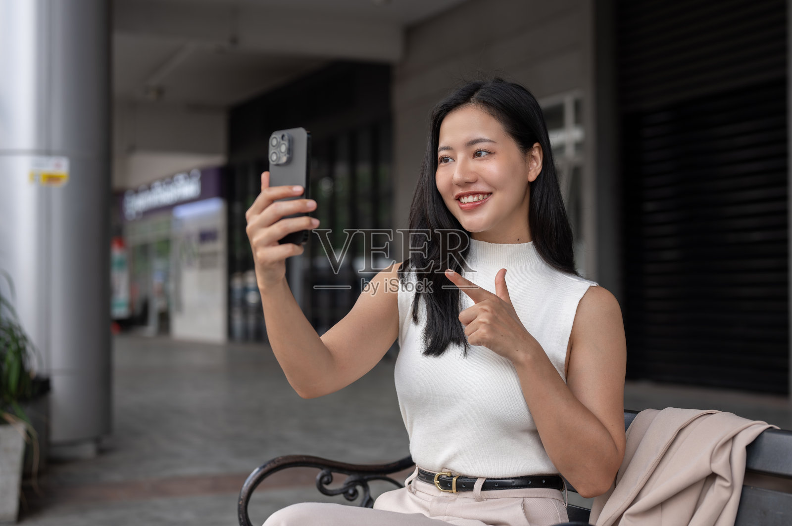 一位美丽的亚洲女性坐在户外的长椅上，用智能手机打视频电话。照片摄影图片