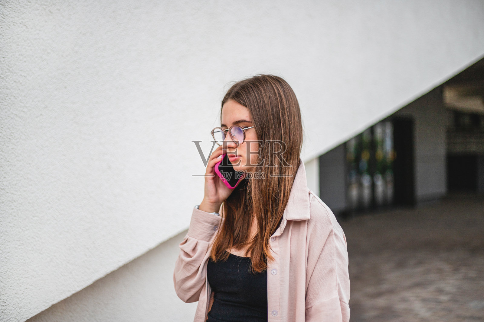 一位戴着眼镜、身穿米色衬衫的年轻漂亮女孩侧身站在一堵白色的墙上，正在讲电话照片摄影图片