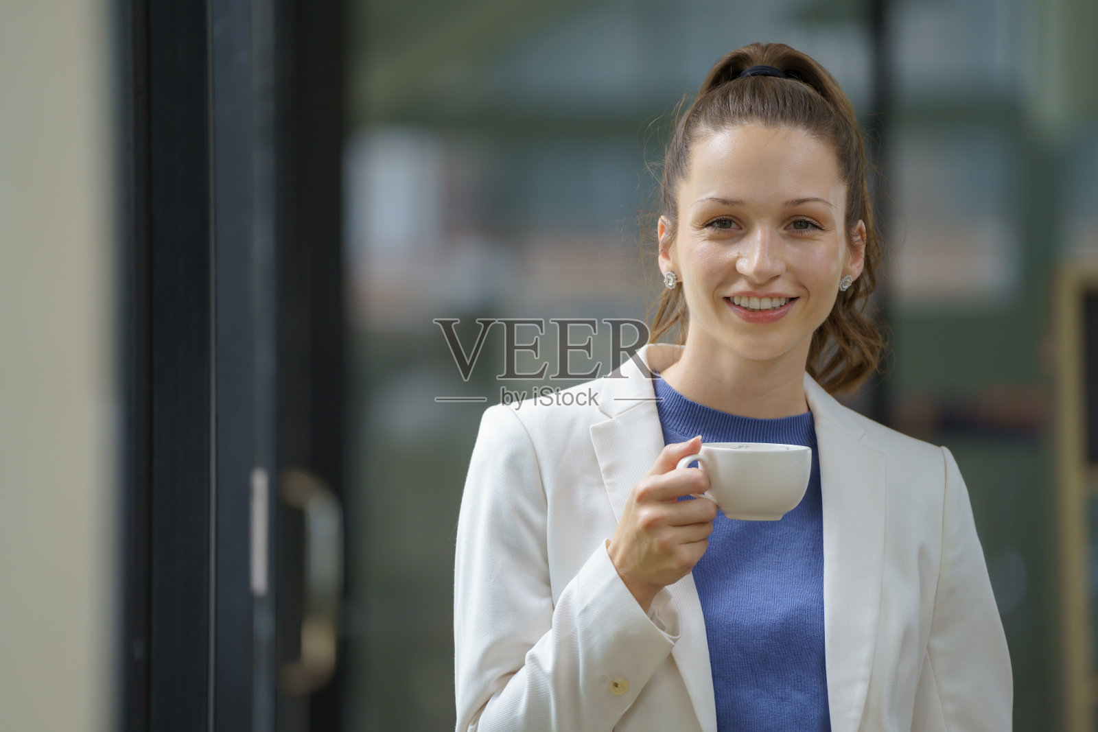 一位女商人站在办公室里，端着一杯咖啡，看着相机。照片摄影图片