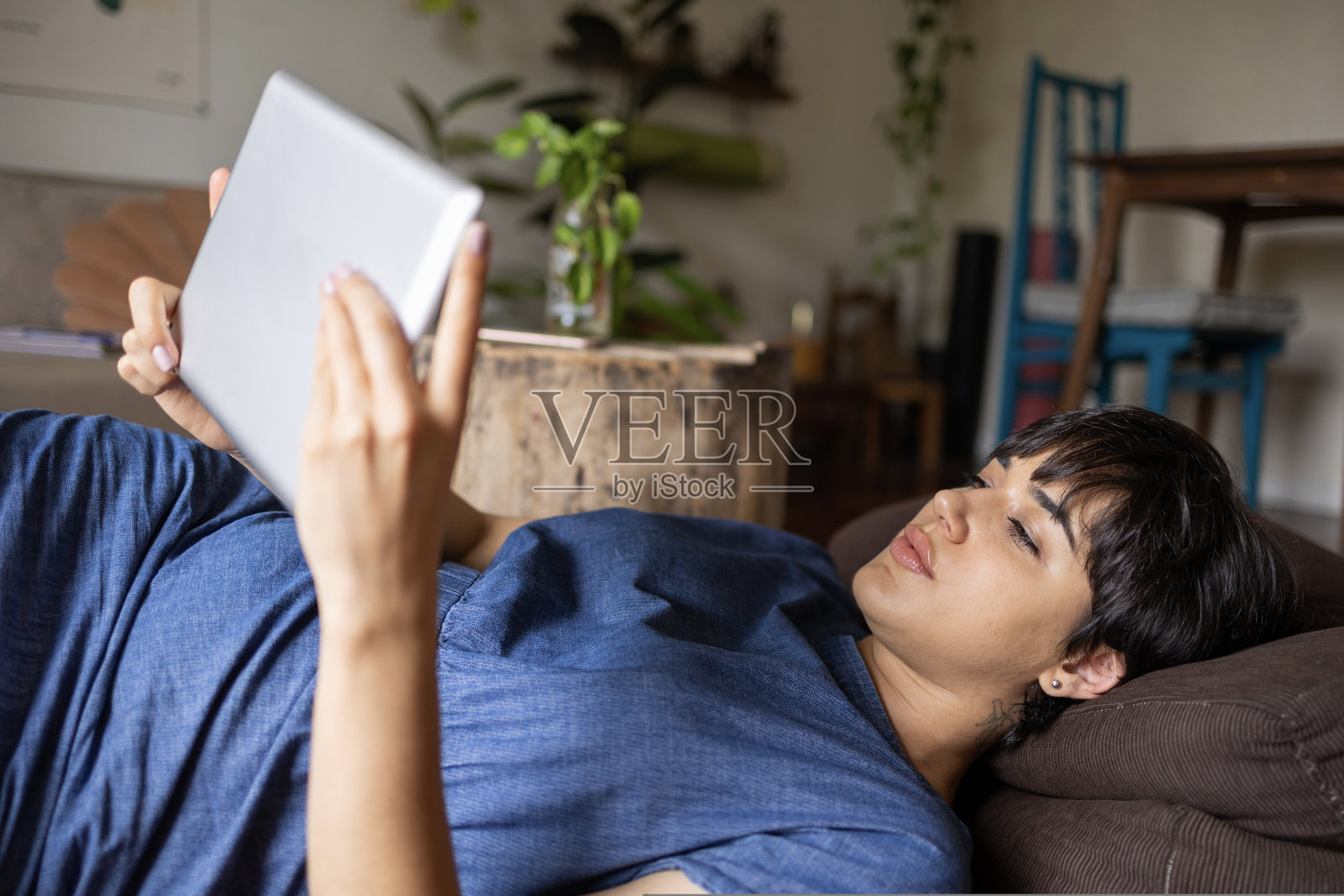 轻松的年轻拉丁女孩，短发，拿着平板电脑。她穿着一件蓝色的裙子躺在满是植物的房间里。照片摄影图片