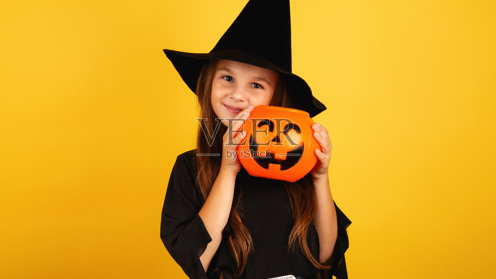 一个长头发的小女孩穿着万圣节的女巫服装微笑着，手里拿着一个黄色背景的南瓜形状的糖果碗。照片摄影图片