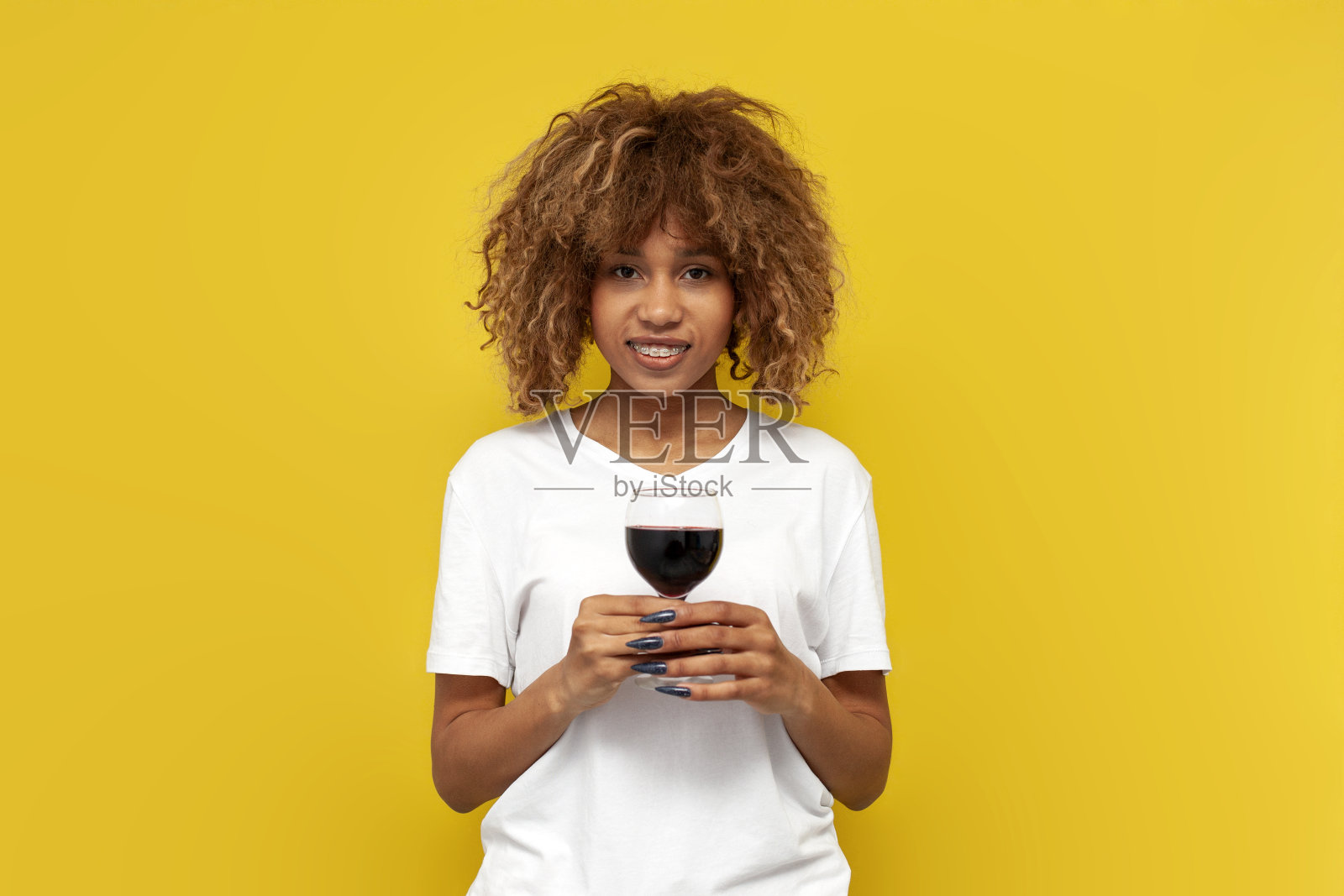 年轻卷曲的美国女孩带着牙套拿着一杯酒在黄色孤立的背景照片摄影图片