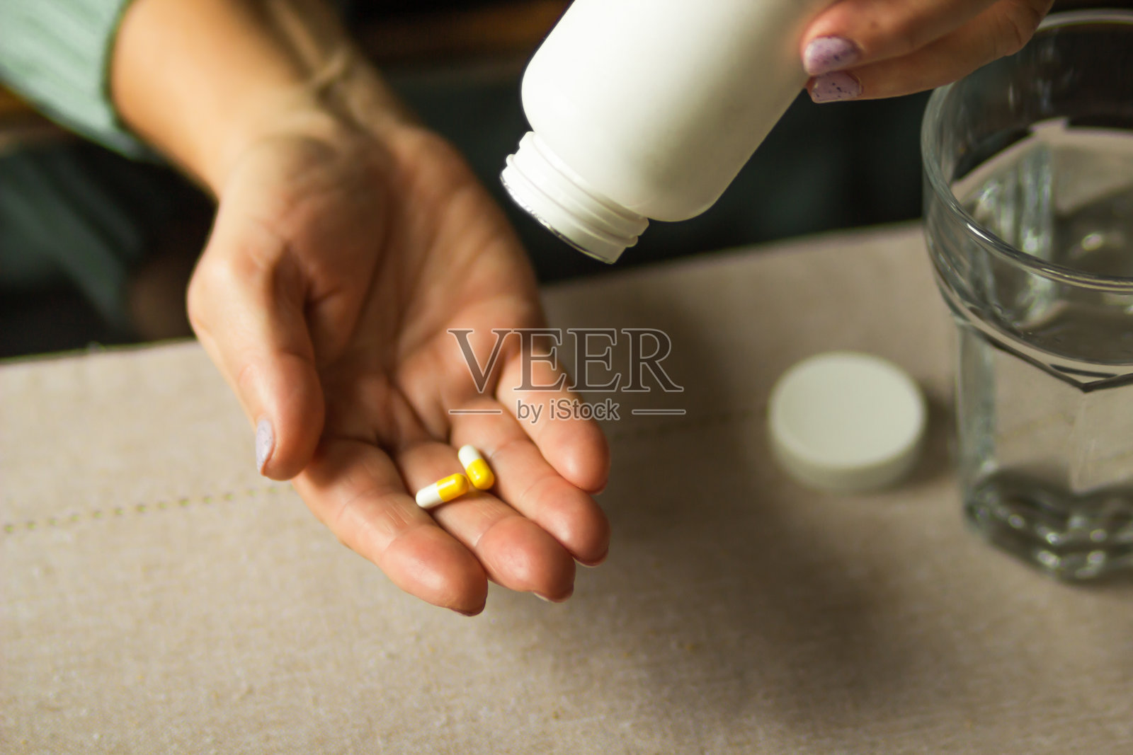 老妇人手里拿着白色的塑料瓶和两粒胶囊，准备用一杯水服用补充药片。照片摄影图片