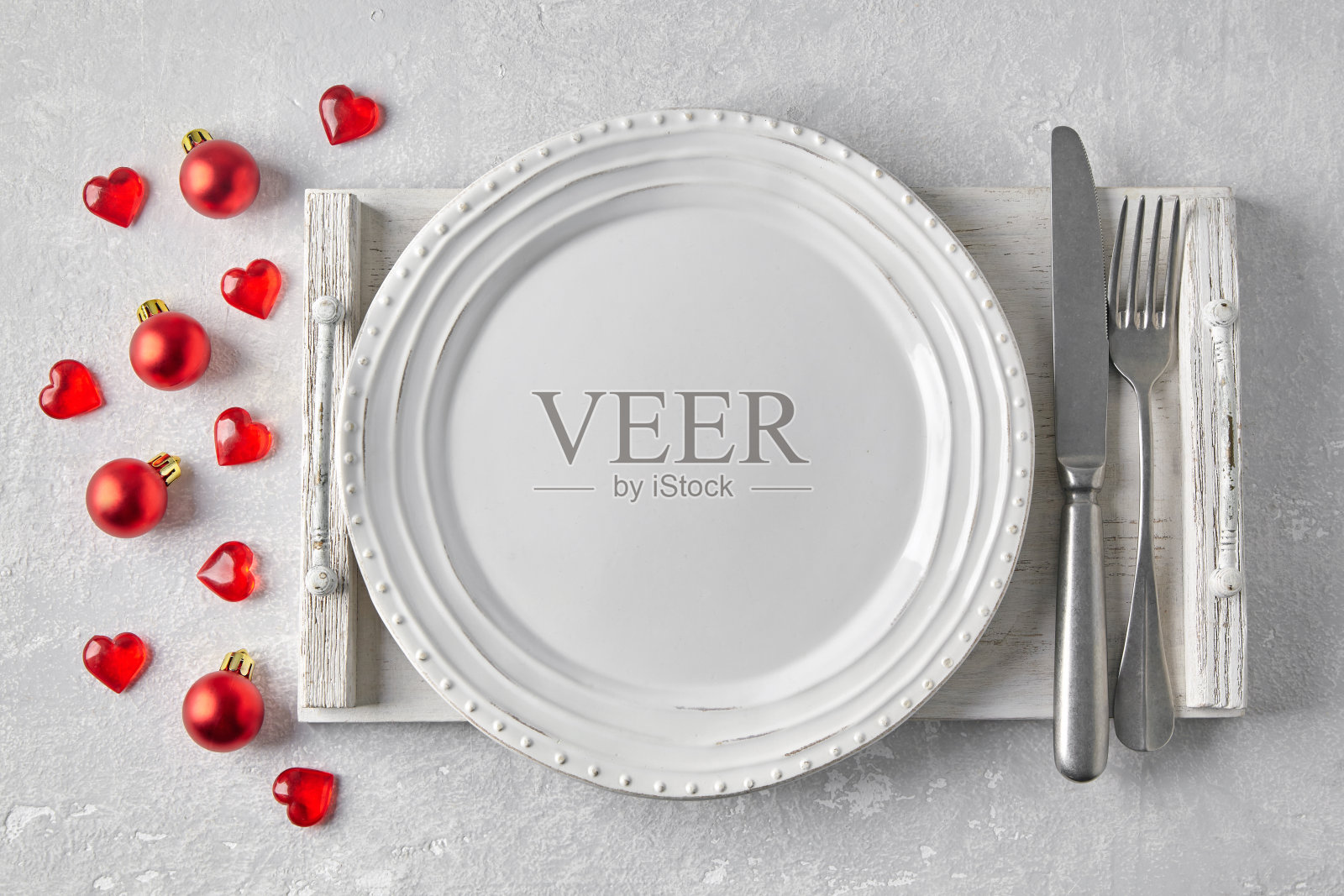 白色木托盘上的白色空盘子和餐具放在灰色混凝土桌子上，周围是红色的圣诞球和心形图案。在菜单上展示食物的模板照片摄影图片