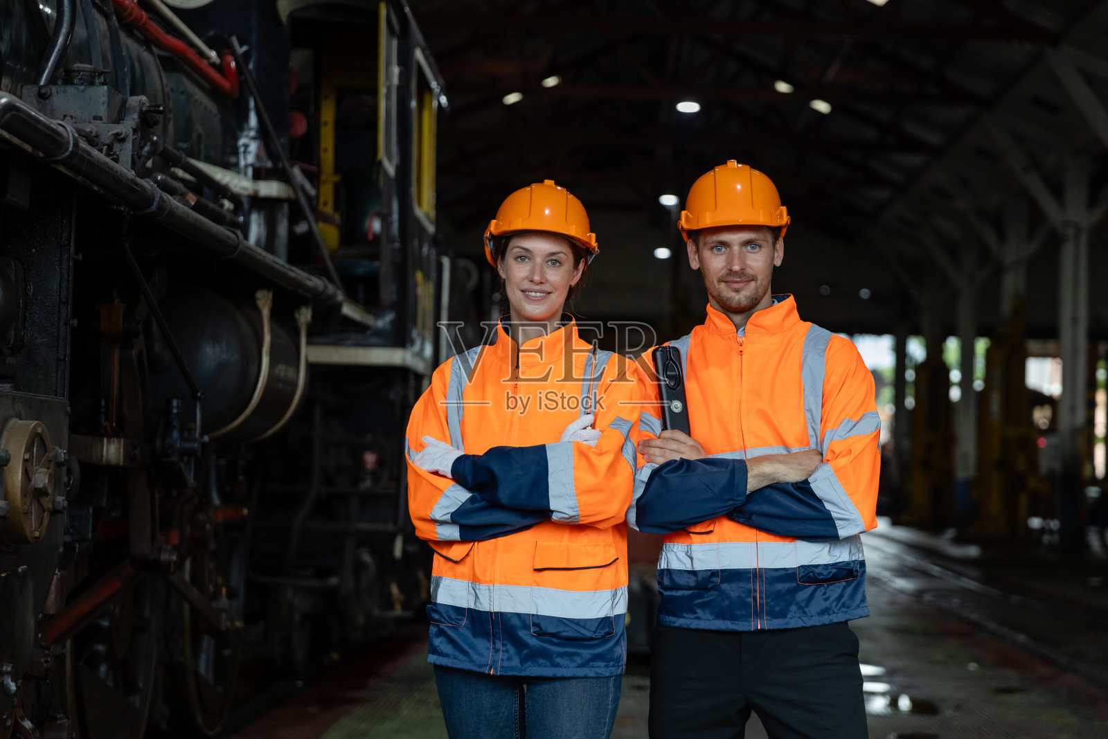 肖像组工程师双手交叉站在维修中心的火车柴油机前。班组铁路工程师检查维修工程列车柴油。照片摄影图片