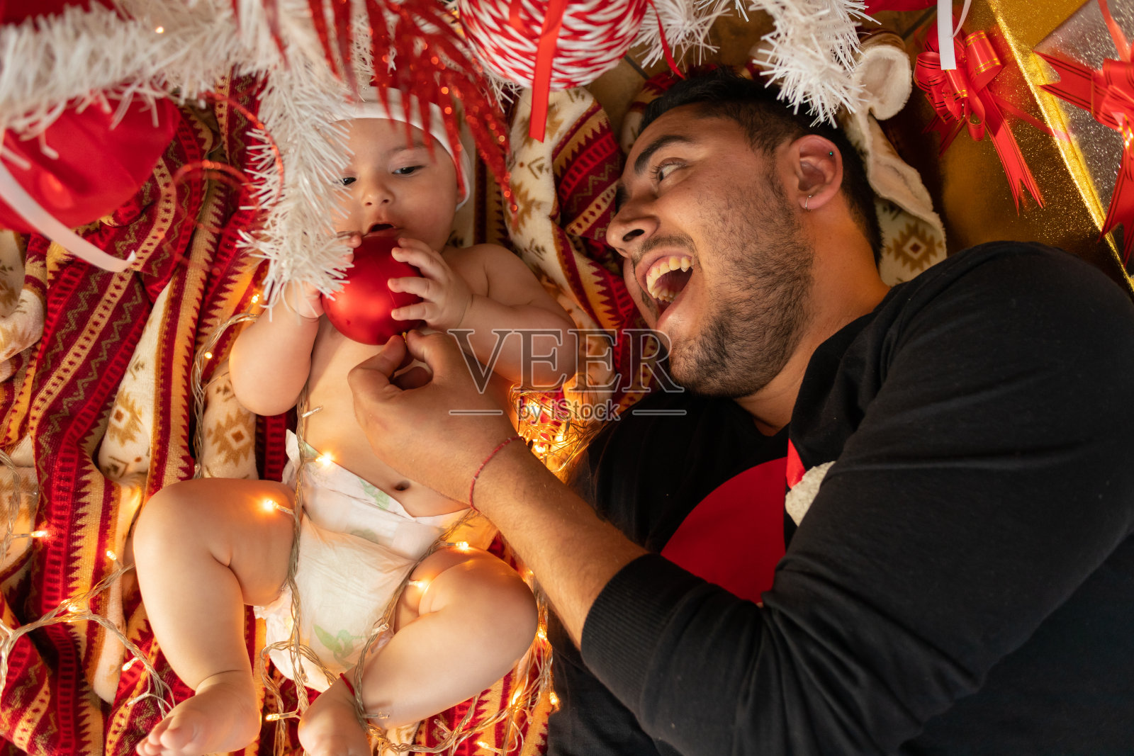 西班牙裔父亲和儿子在圣诞树下笑着玩球和灯照片摄影图片
