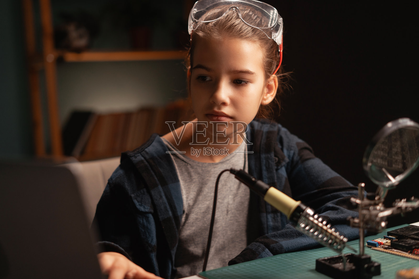 聪明的年轻女孩正在研究焊丝和电路板，看着笔记本电脑在她的科学爱好项目中工作。教育照片摄影图片