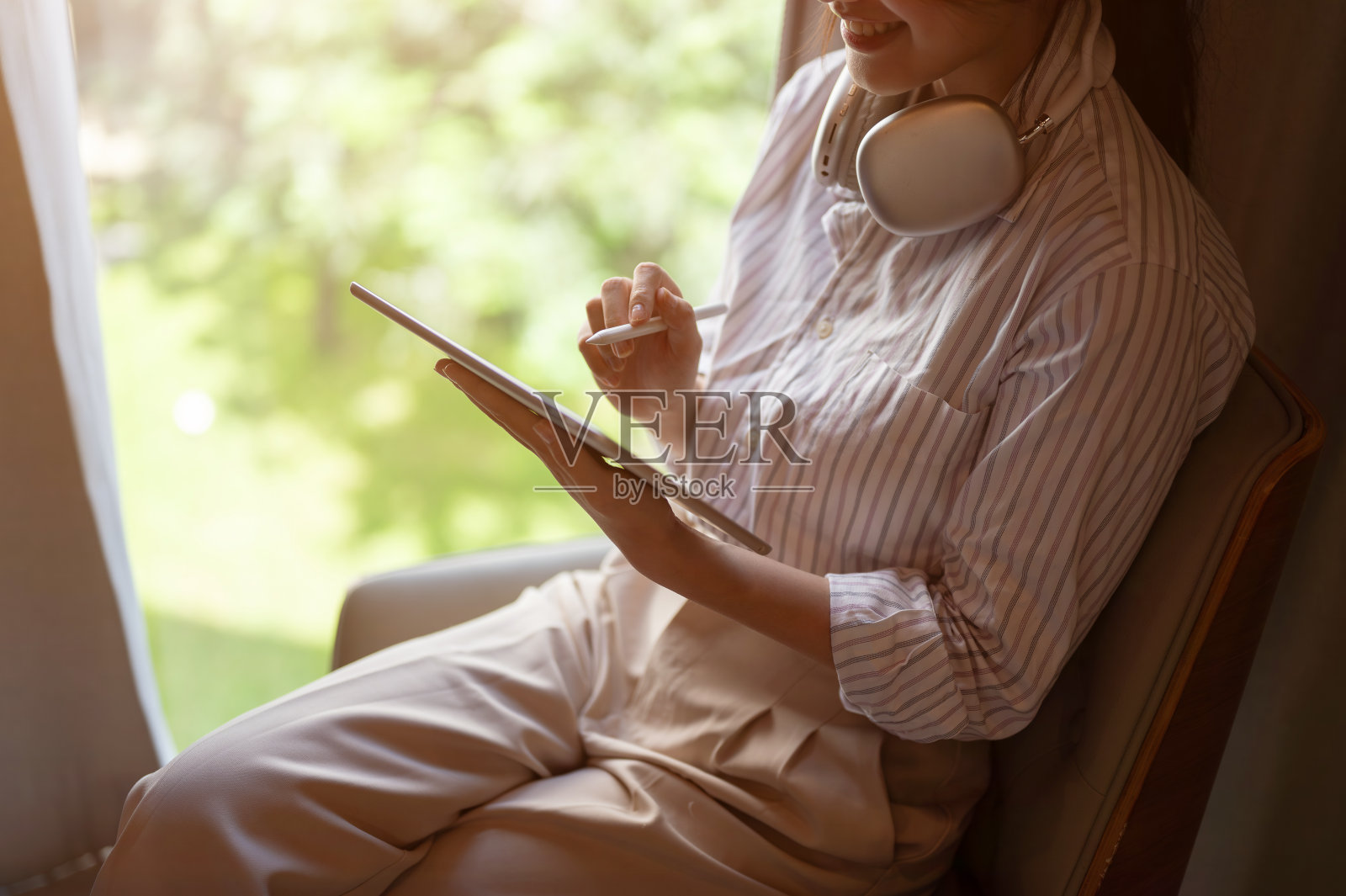 一位快乐的亚洲女性坐在室内靠窗的位置上使用她的数码平板电脑。照片摄影图片
