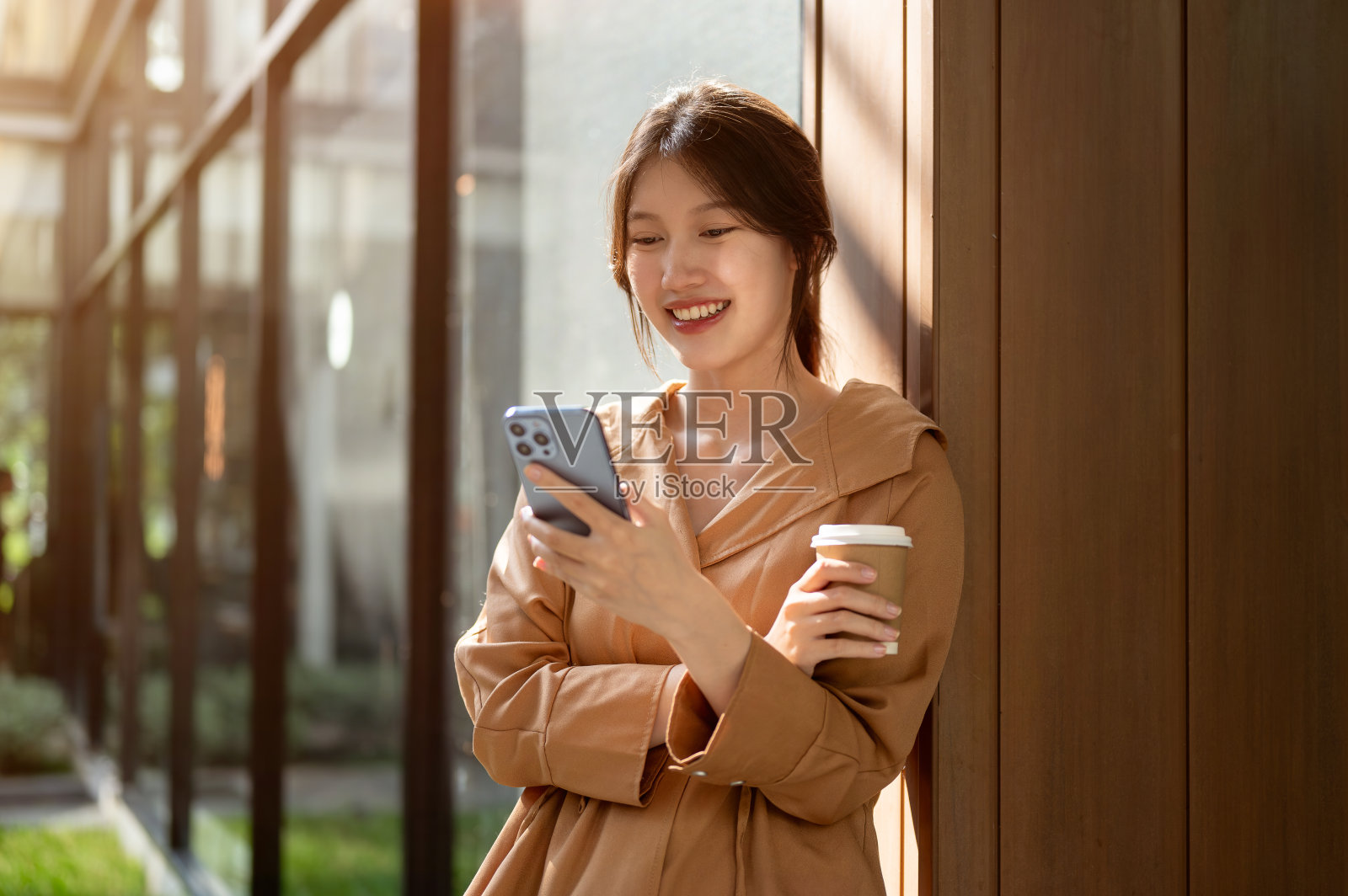 一位迷人的亚洲女性倚在户外的墙上，一边用智能手机一边喝着咖啡。照片摄影图片