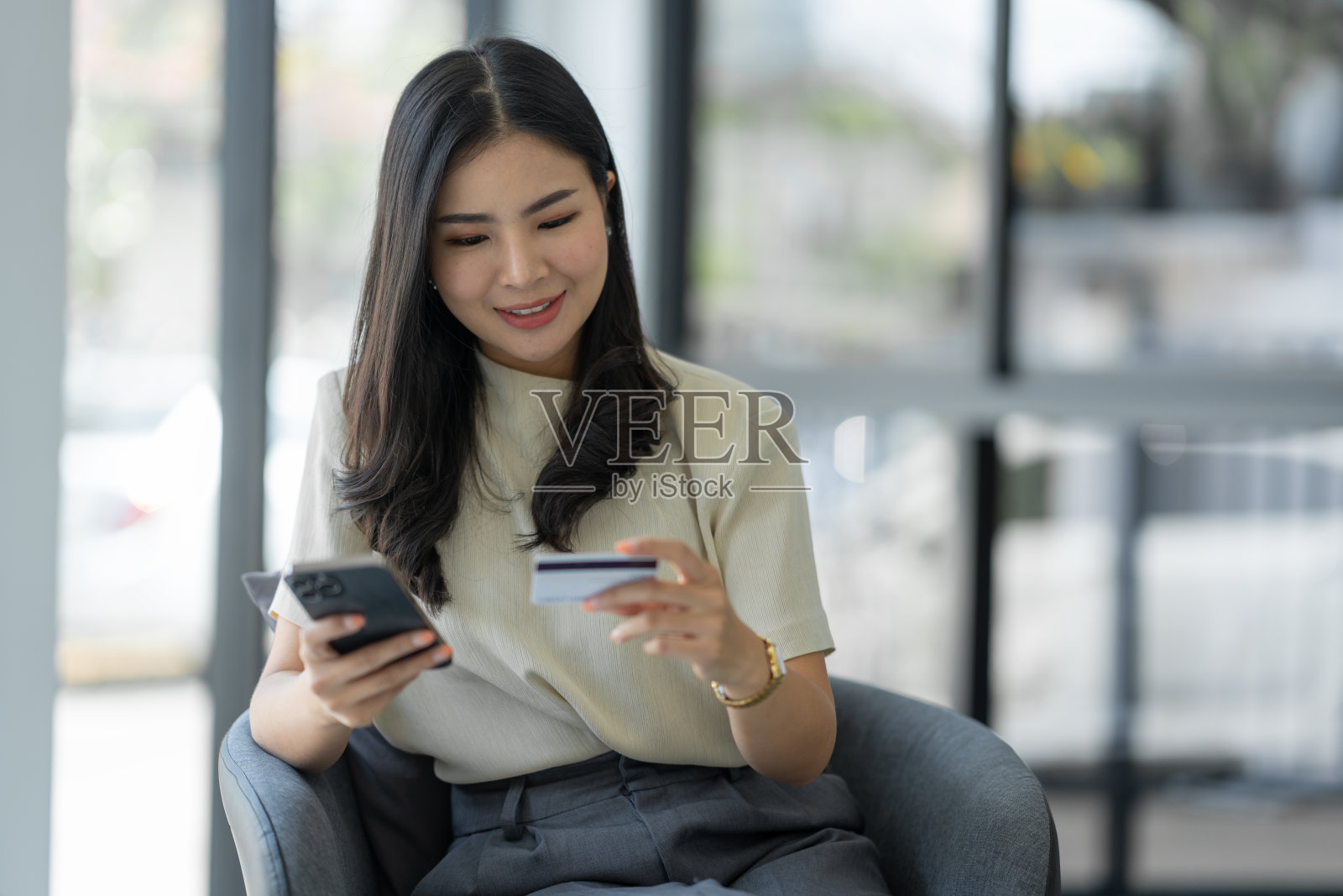 自信的亚洲年轻女子坐在办公室的沙发椅上，用手机打开应用程序，用信用卡进行网上购物。照片摄影图片