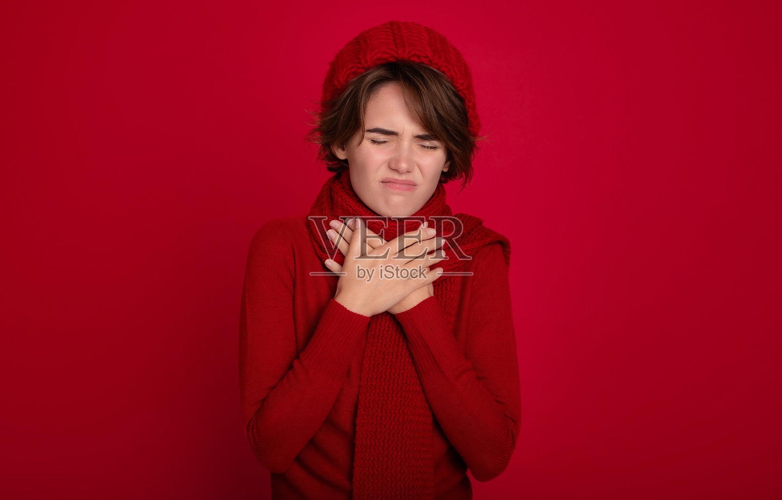 特写:年轻悲伤的女人穿着红色毛衣，围巾和帽子遮住嘴，双手打喷嚏咳嗽，孤立在工作室的红色背景。健康、病、病、病、治、感冒、季节流感的概念照片摄影图片