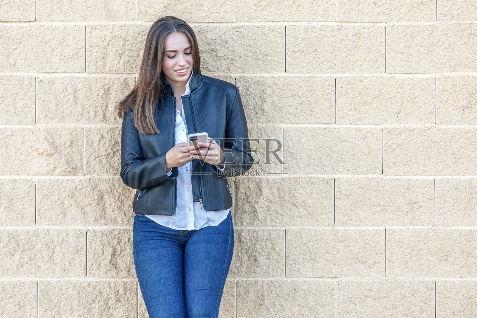 一个拿着智能手机微笑着站在砖墙上的女人照片摄影图片