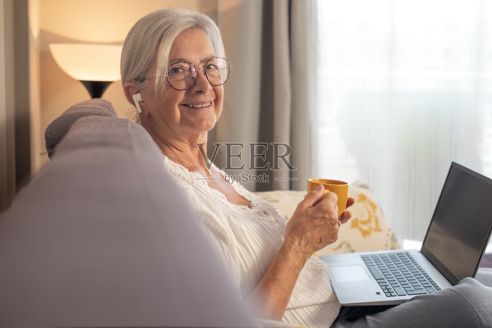 轻松帅气的资深女性坐在沙发上拿着咖啡杯看着相机，使用笔记本电脑技术，戴着耳机听着，享受着无忧无虑的时光照片摄影图片