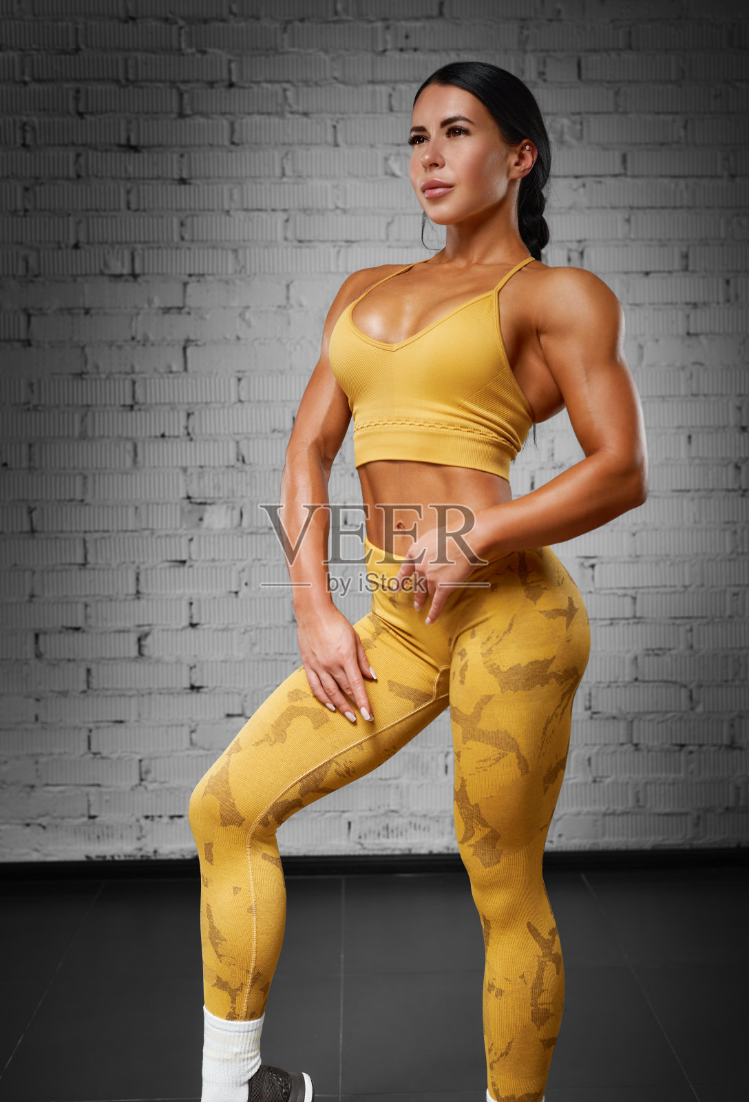 性感健身的女人。一个漂亮的运动女孩在工作室里穿着黄色的训练服，背景是一堵砖墙照片摄影图片