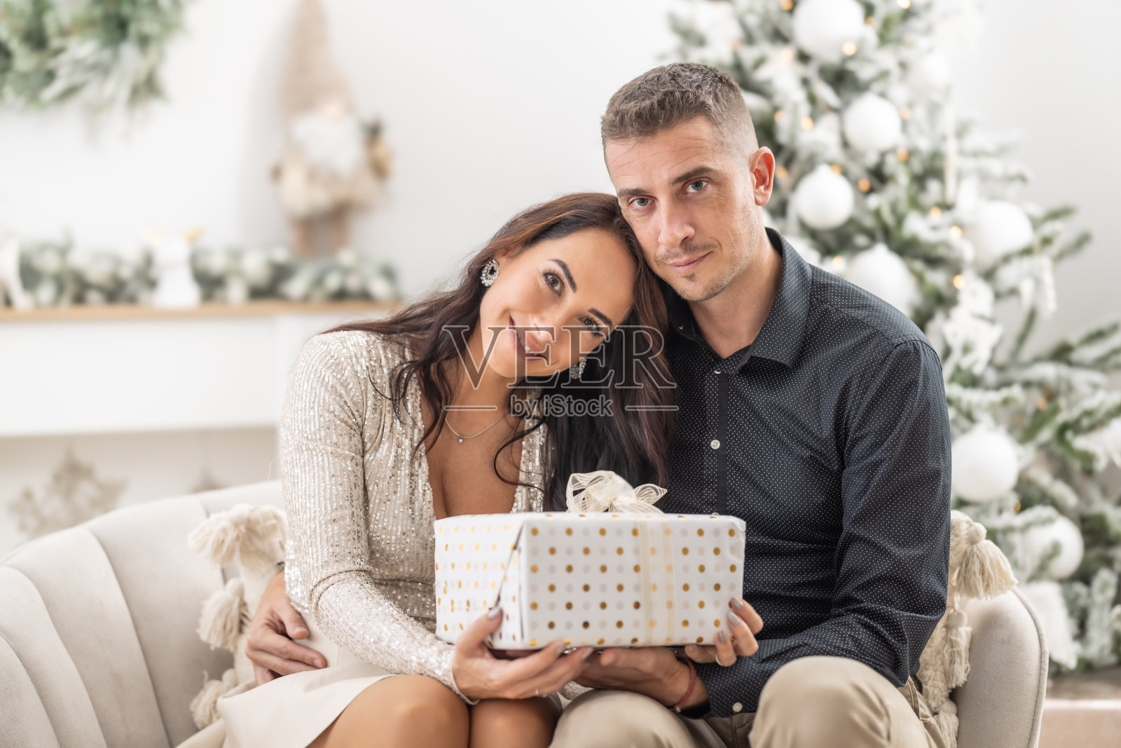 一对相爱的情侣坐在圣诞树前的沙发上，手里拿着圣诞礼物。照片摄影图片