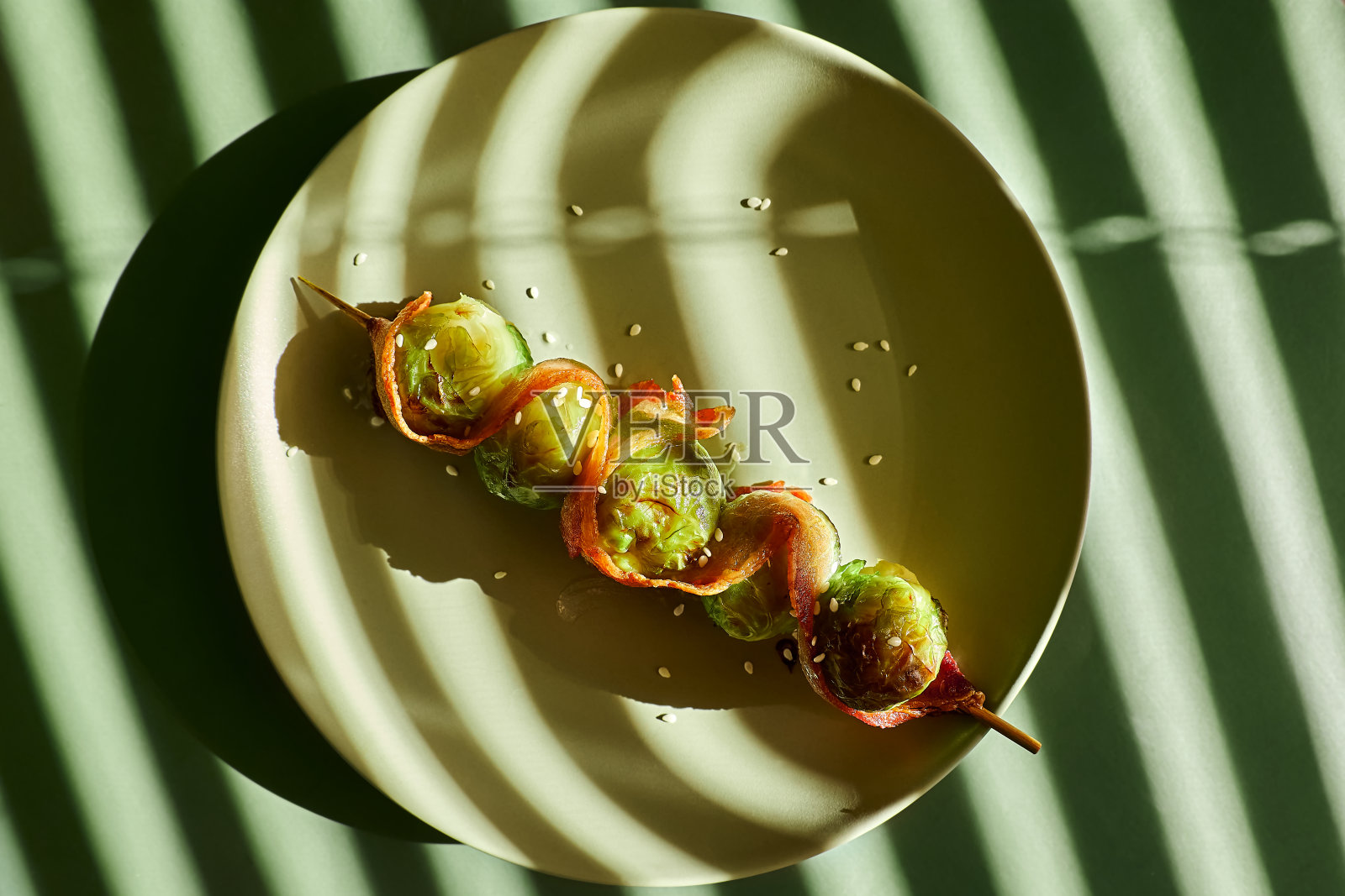 用培根包裹的布鲁塞尔芽串，烤好后放在绿色的盘子上，绿色的背景上有几何形状的硬阴影。照片摄影图片