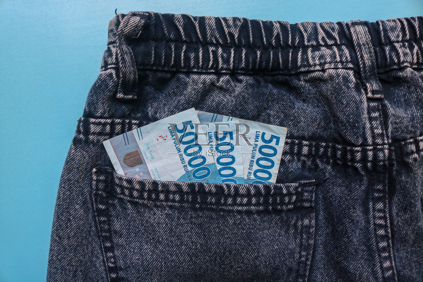 把钱放在灰色牛仔裤的后口袋里，印尼盾的钱放在牛仔裤口袋里，在蓝色的背景上隔离开来照片摄影图片