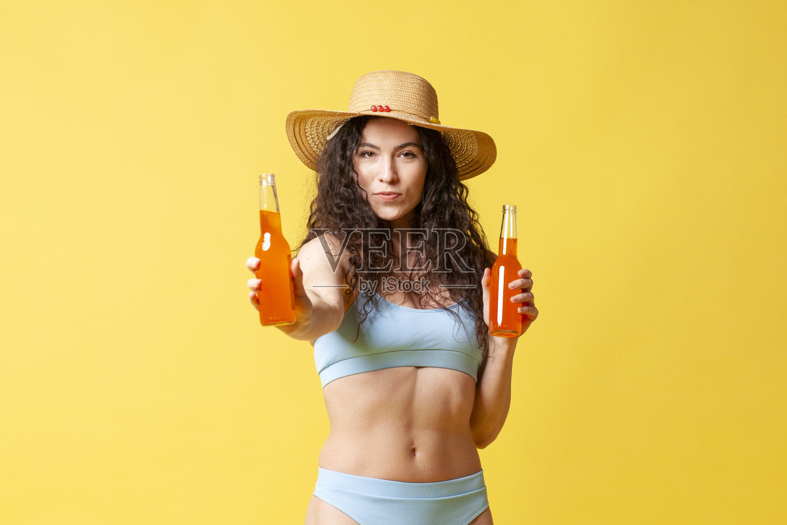 身穿蓝色泳衣的黑发女孩拿着装有橙色饮料的瓶子，并在黄色背景上提供饮料照片摄影图片
