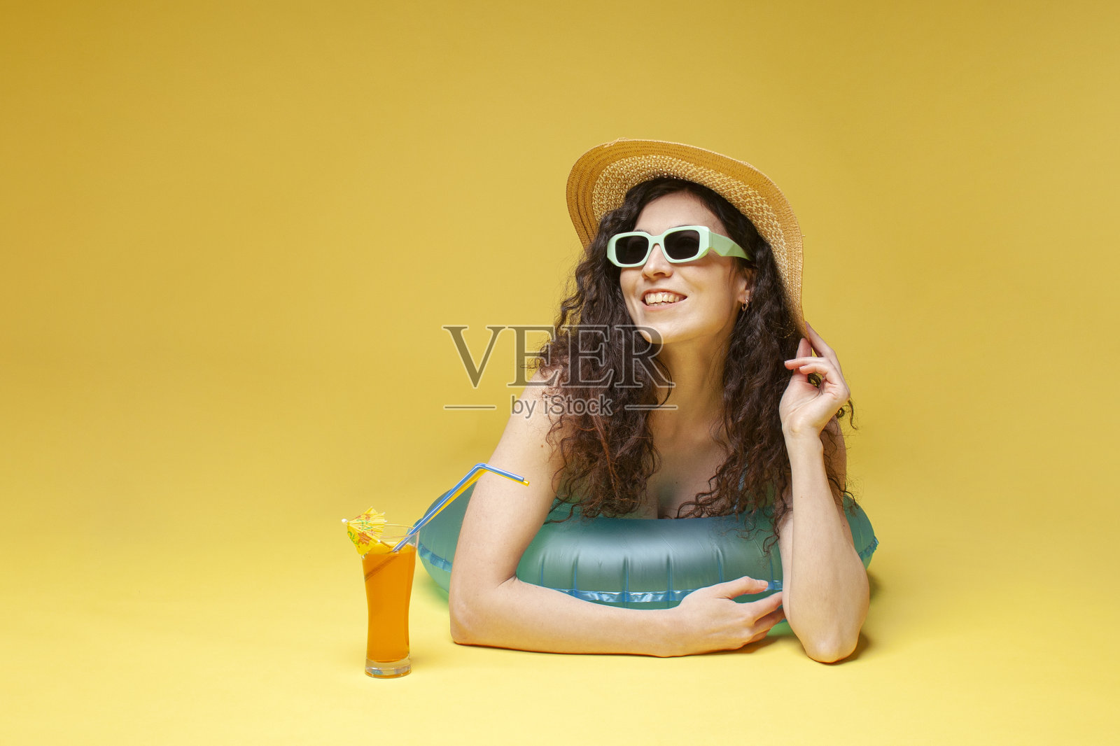 戴草帽的年轻女孩躺在一个充气游泳圈上，黄色背景上有一杯橙色鸡尾酒照片摄影图片