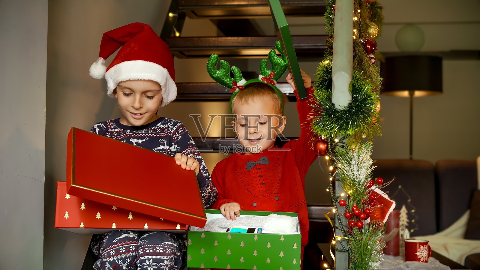 两个快乐的男孩穿着睡衣，戴着圣诞老人的帽子，坐在木楼梯上打开圣诞礼物。寒假的家庭庆祝活动。照片摄影图片