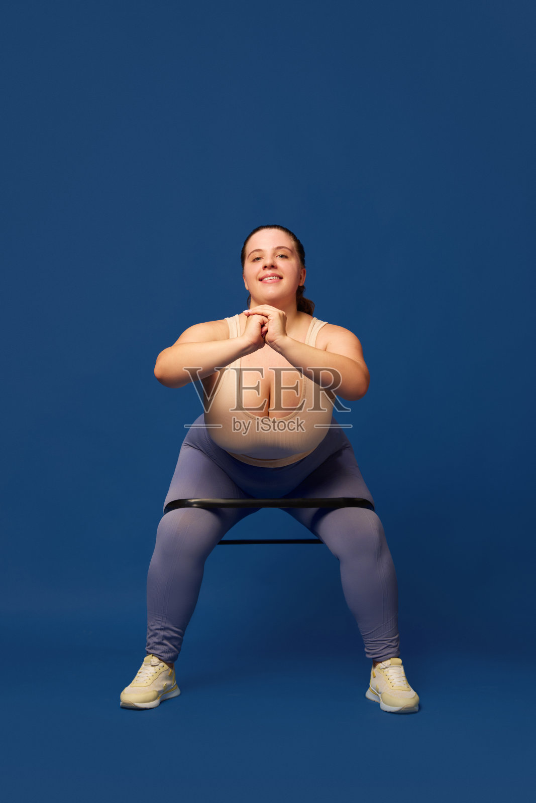 年轻的超重女性用健身松紧带训练，在蓝色的工作室背景下做深蹲。运动,body-positivity照片摄影图片