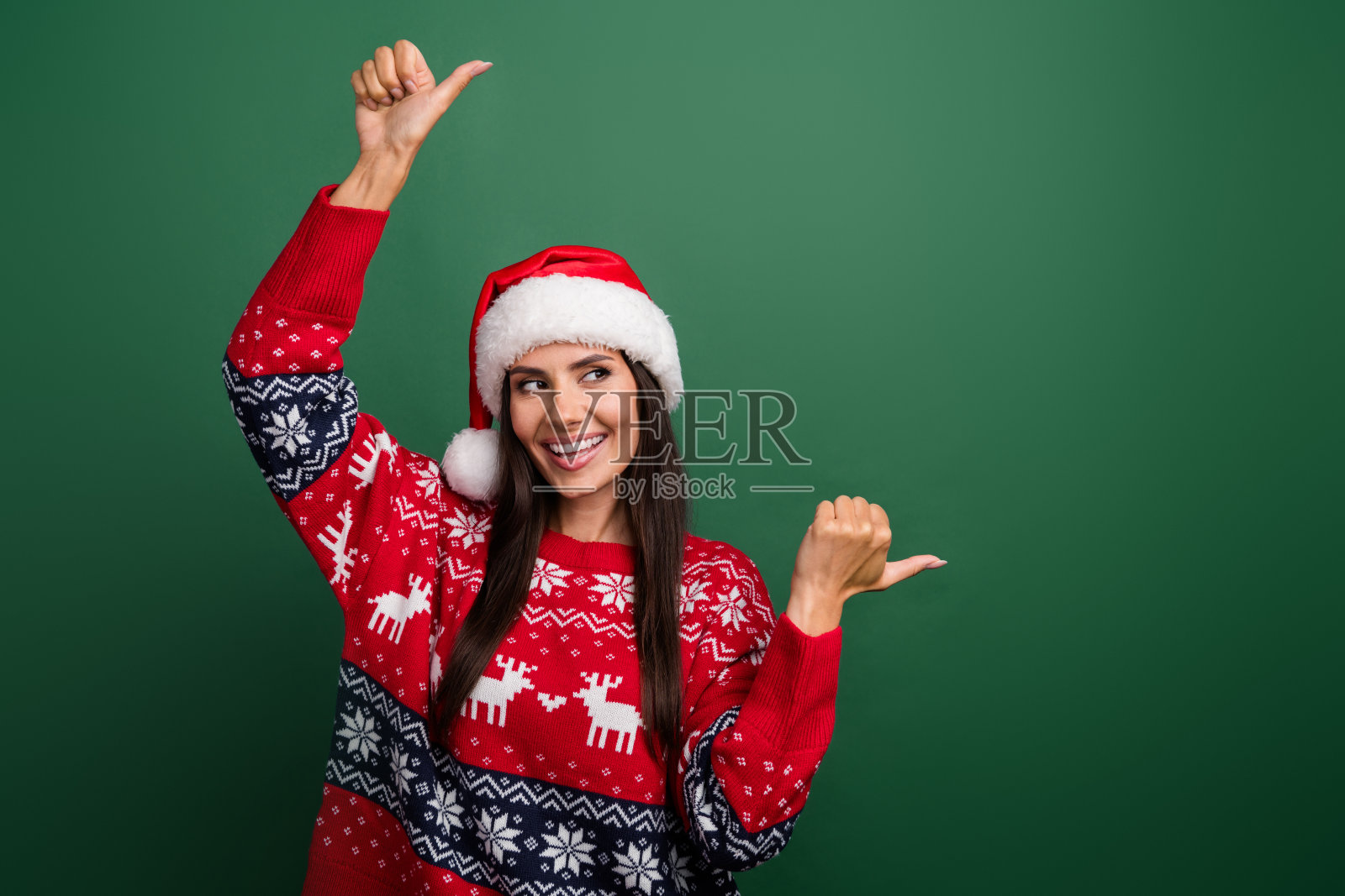 梦幻可爱的圣诞女助手穿着印花圣诞毛衣的照片，显示两个拇指空白的空间孤立的绿色背景照片摄影图片