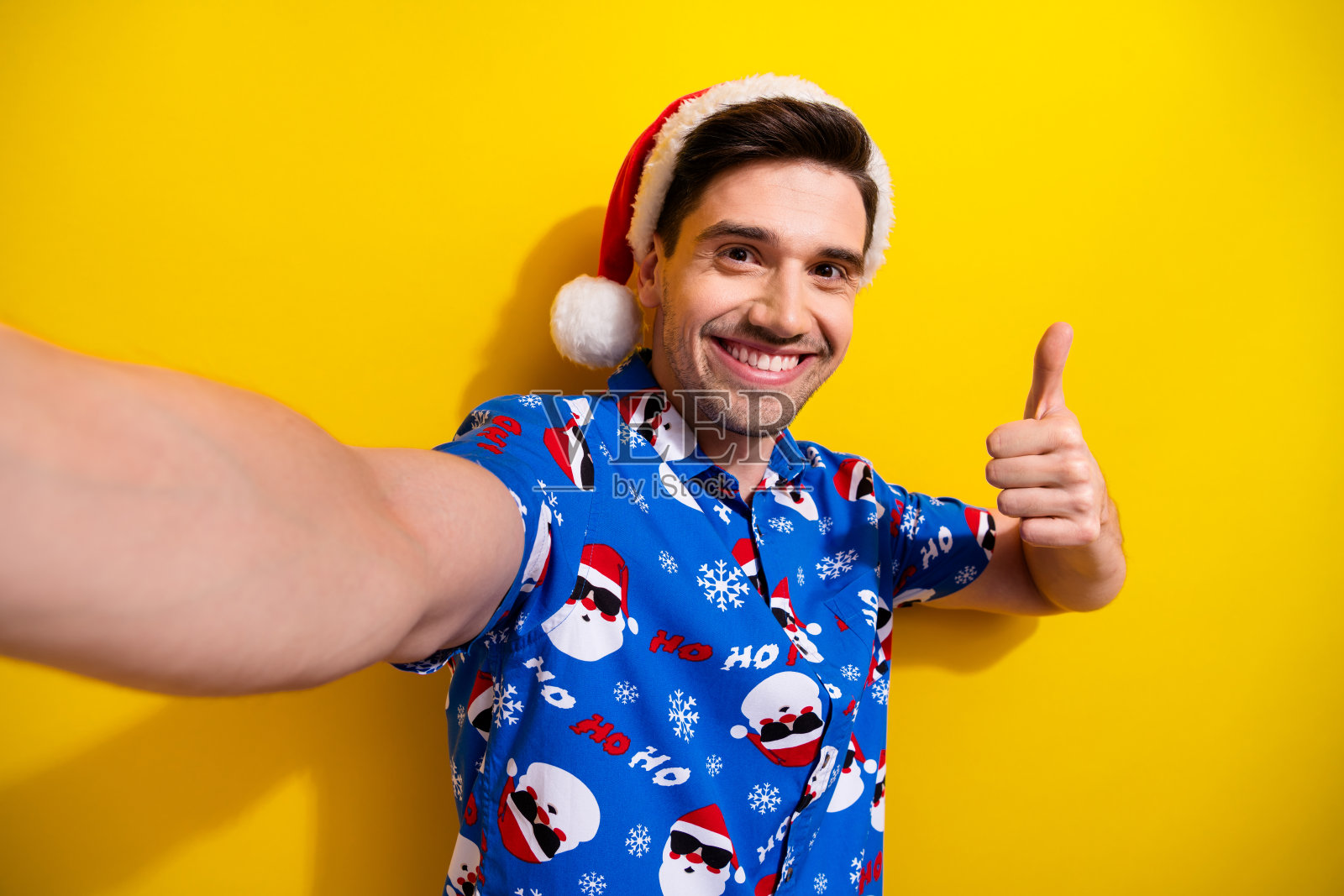 新视频博客小伙戴着泡泡帽的自拍，展示了蓝色衬衫，圣诞老人的印花，就像反馈一样孤立在黄色背景上照片摄影图片