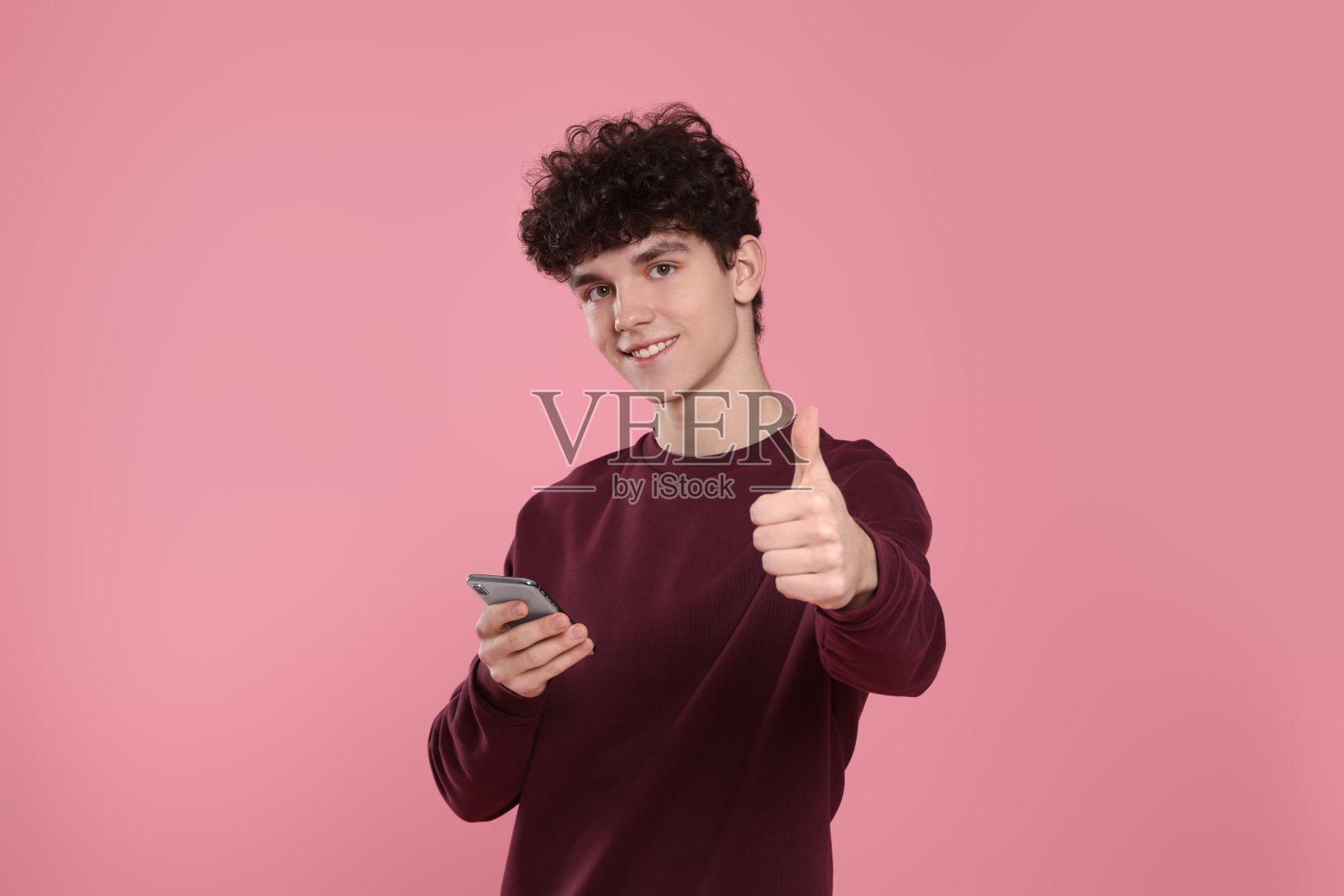 一个微笑的十几岁男孩的肖像，他的智能手机在粉红色的背景上竖起大拇指照片摄影图片