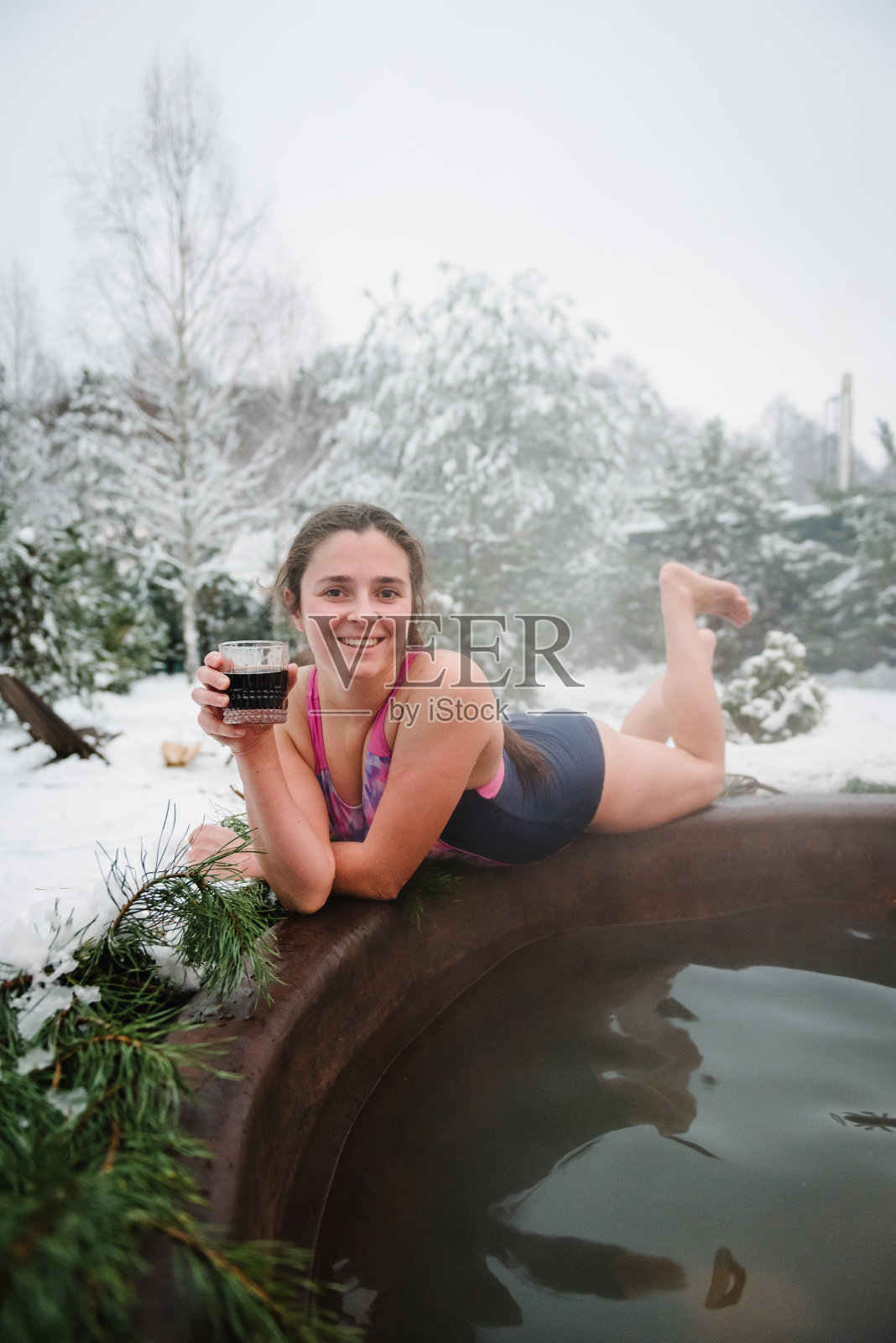 在白雪皑皑的森林里享受温泉浴的女性。山区寒假，热水理疗概念。下雪时，年轻的女孩在户外。女人喝冷饮，在户外洗热水澡放松。照片摄影图片