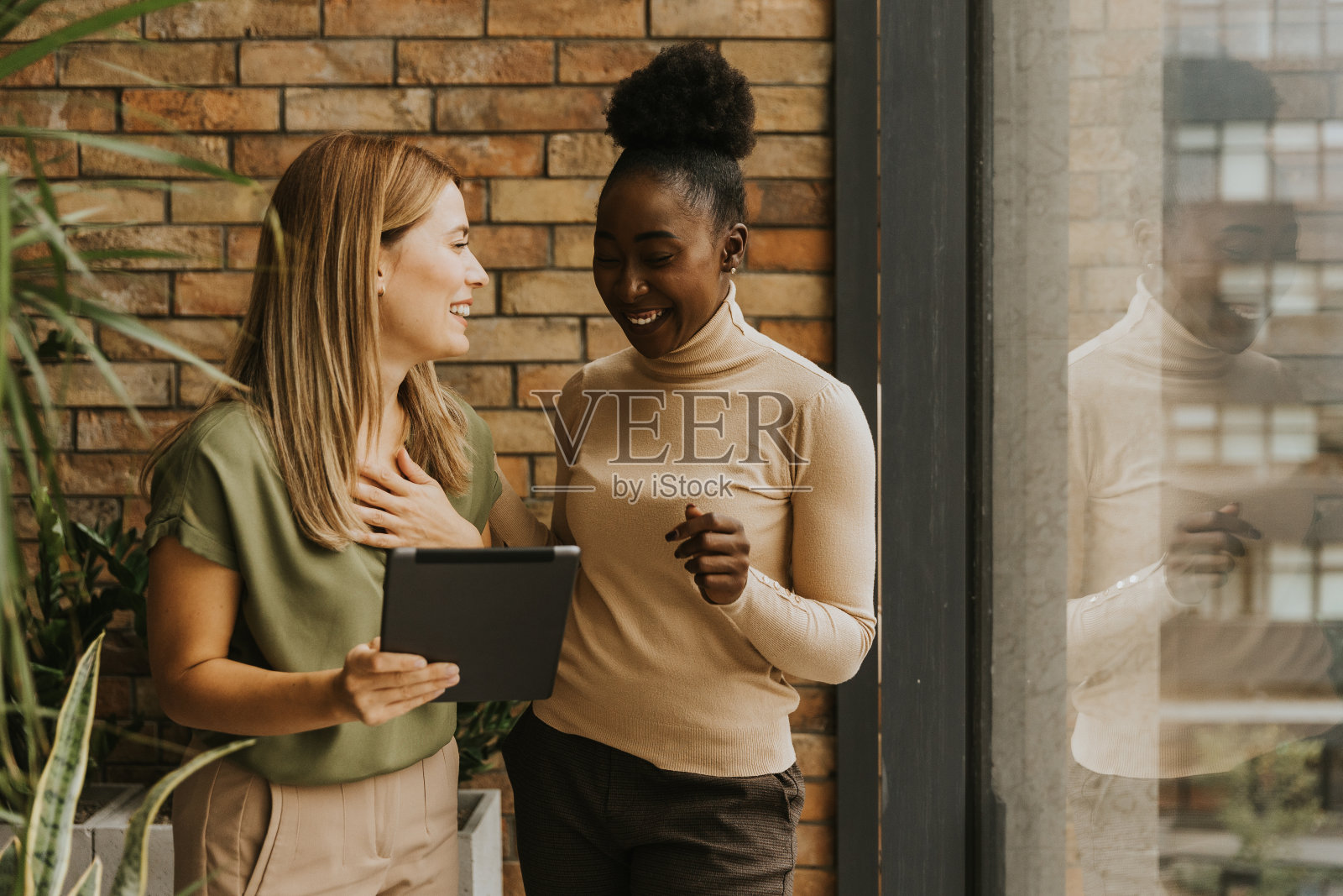在工业风格的办公室里，两个拿着数码平板电脑的年轻女商人站在砖墙旁照片摄影图片