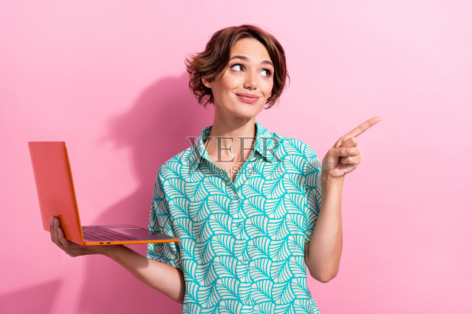 迷人的女孩拿着笔记本电脑的照片，看着手指空白的空间，在粉红色的背景上提供孤立的广告照片摄影图片