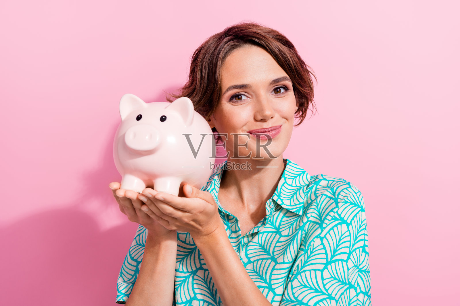 漂亮积极的年轻女孩的肖像手臂拿着钱储蓄银行猪隔离在粉红色的背景照片摄影图片