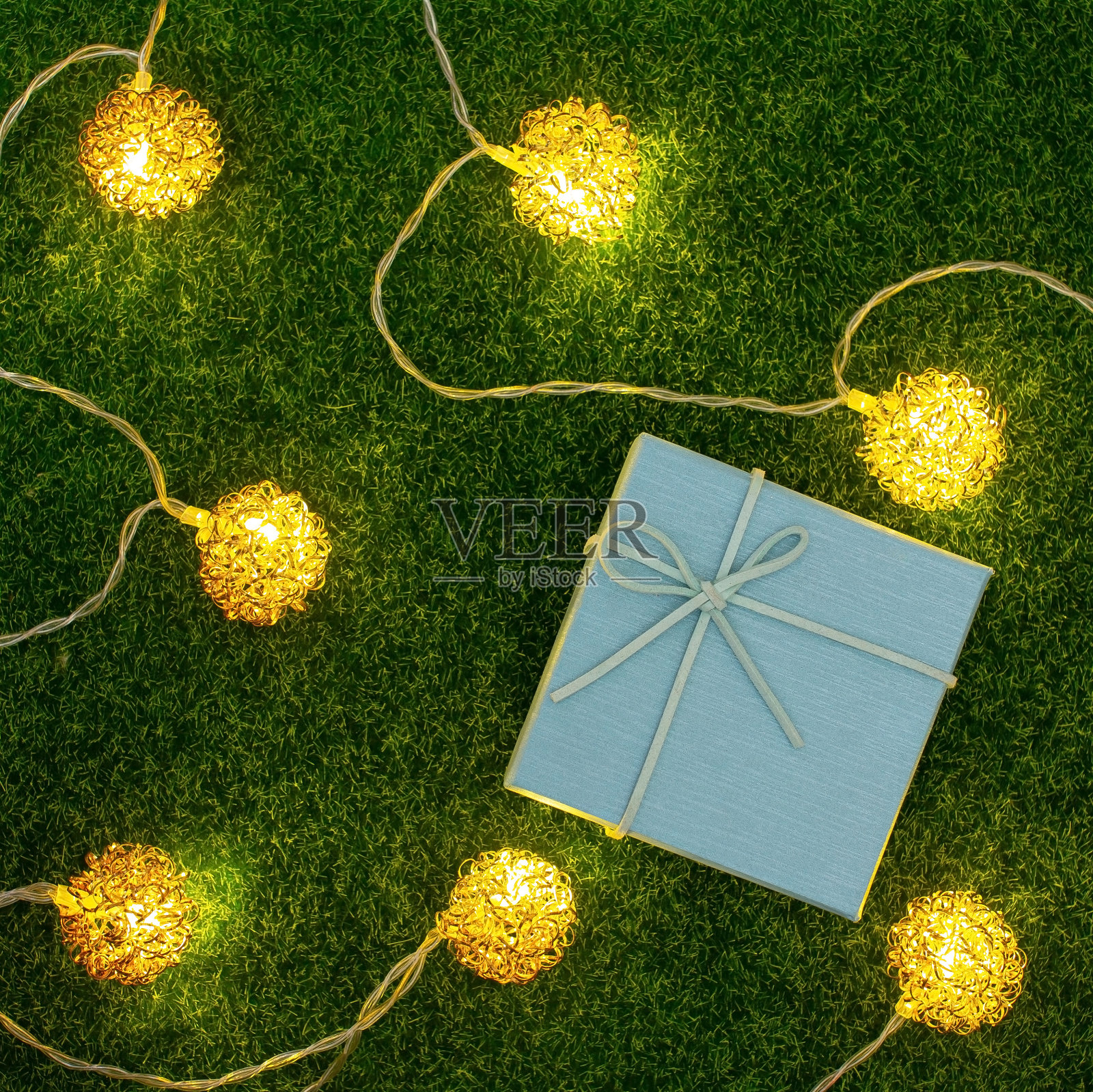一个蓝色的礼品盒放在绿色的人造草坪上，周围有装饰灯。照片摄影图片