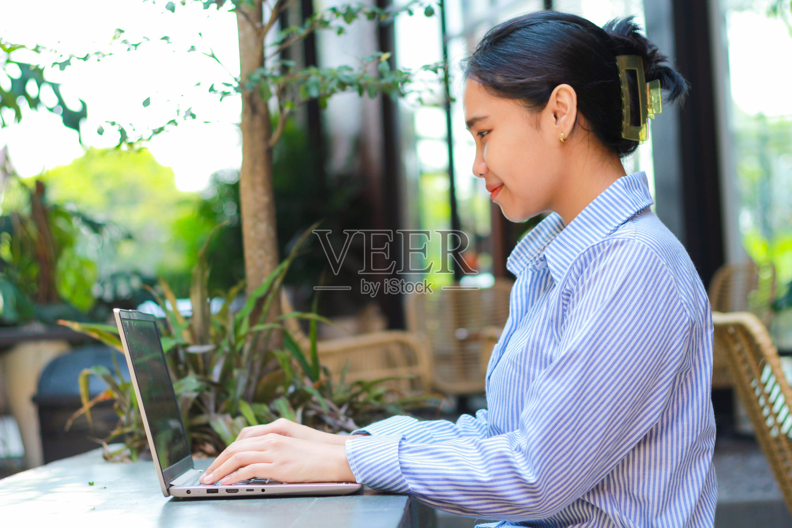 快乐的亚洲女人坐在户外咖啡馆里远程用笔记本电脑工作，穿着蓝色条纹衬衫，背景是绿色植物照片摄影图片