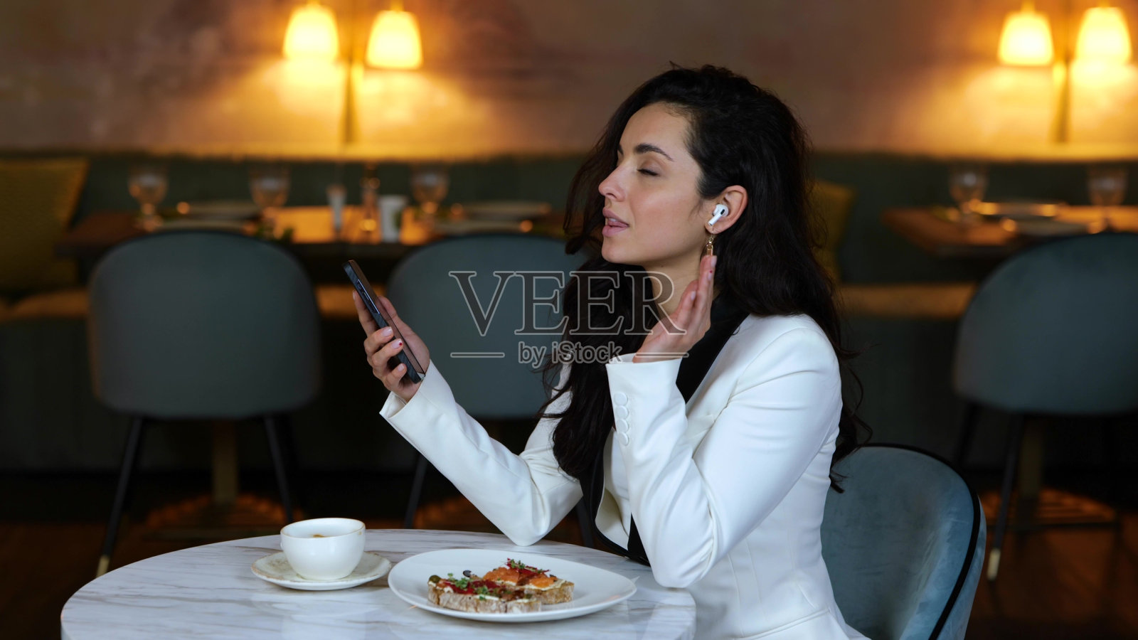 快乐的年轻女士坐在餐厅里拿着智能手机，戴着无线耳机。唱歌的女人。商业，生活理念。慢动作照片摄影图片