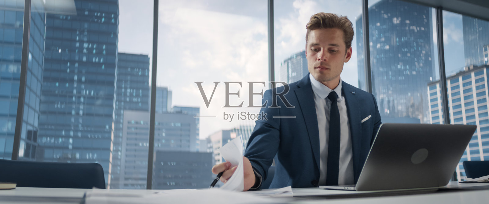西装革履的自信商人坐在现代办公室的办公桌前，使用笔记本电脑，旁边的窗户可以看到大城市的景色。成功的财务经理思考工作项目。照片摄影图片