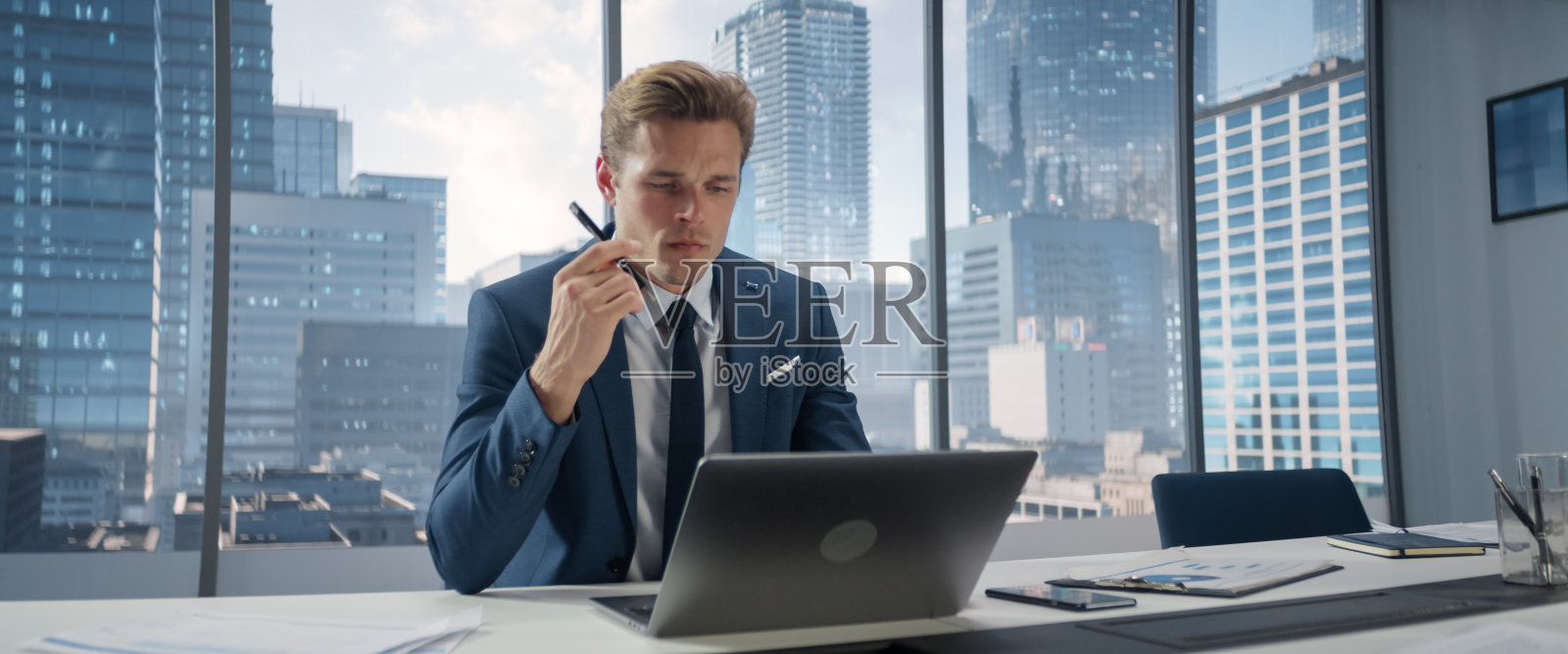 西装革履的自信商人坐在现代办公室的办公桌前，使用笔记本电脑，旁边的窗户可以看到大城市的景色。成功的财务经理思考工作项目。照片摄影图片