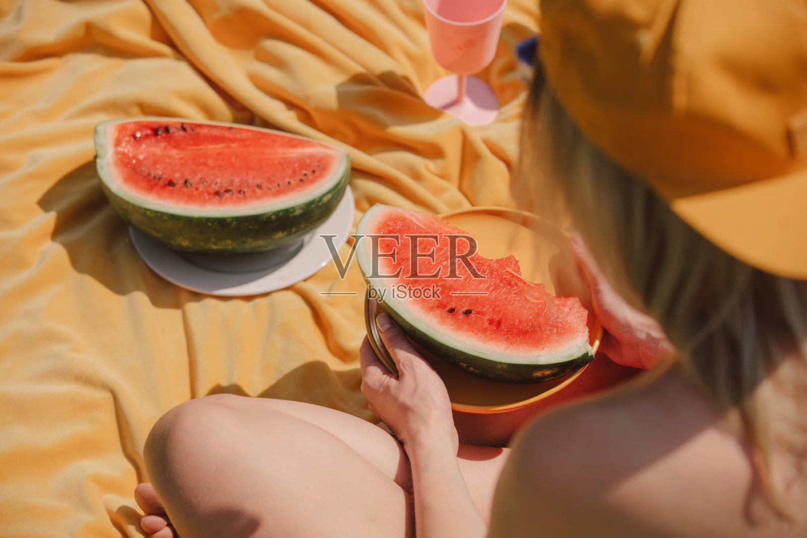 戴帽子的女人坐在黄布上吃西瓜。后视图照片摄影图片